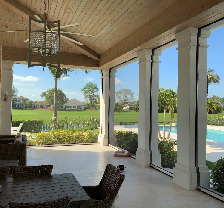 View of a backyard with a swimming pool, palm trees, and a golf course beyond a screened porch with wicker chairs and a wooden table.