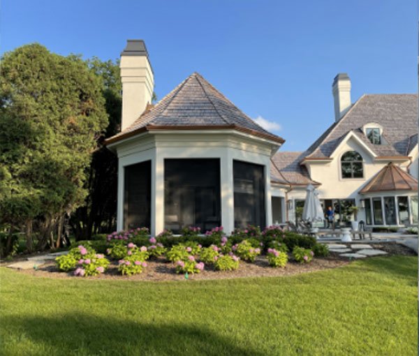 A large Victorian-style house with a turret, front porch, and well-maintained garden with shrubs and flowers, under a bright blue sky.
