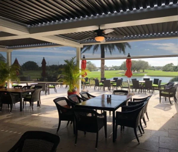 Outdoor dining area with black tables and chairs, red umbrellas, potted plants, and a view of a golf course with trees and a pond under a covered patio.