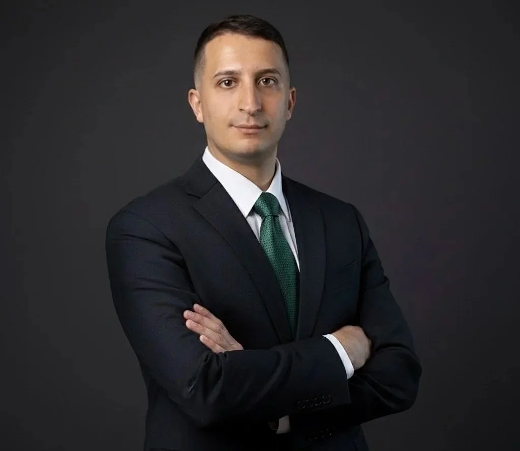 A professional man in a suit with a white shirt and green tie, standing with arms crossed against a dark gray background.
