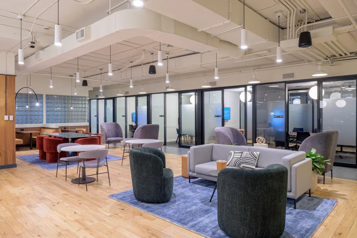 Modern office lounge area with various chairs, sofas, and tables on wooden floors, separated by glass partitions, with ceiling lights and exposed ductwork.