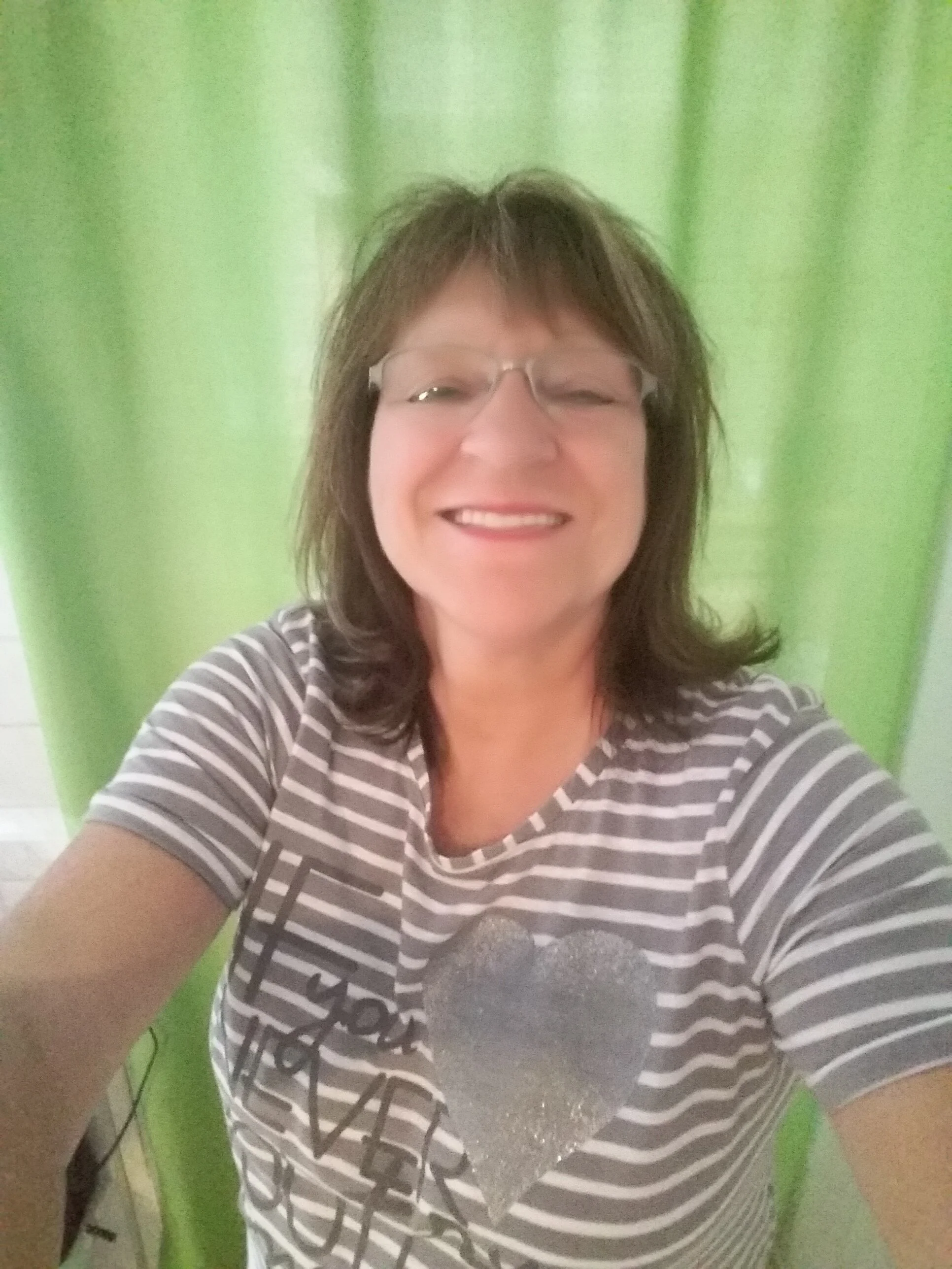 Eine lachende Frau mit Brille vor hellgrüner Kulisse, trägt ein grau-weiß gestreiftes T-Shirt mit einem großen, glänzenden Herzen auf der Vorderseite.