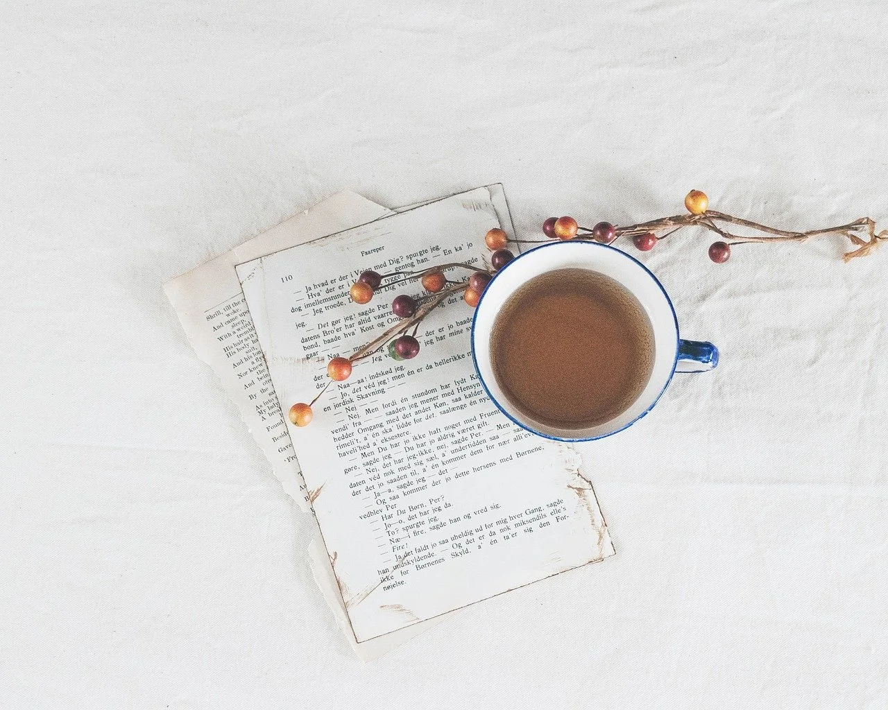 Auf einem weißen Untergrund liegt ein Buch, eine Tasse Kaffee und eine dünne Zweig mit roten Beeren.