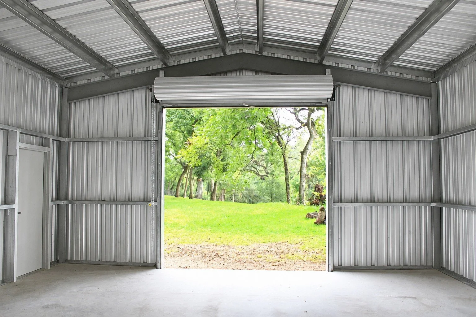 Interior view showing the weld-up steel framing, galvanized roof and wall purlins, metal wall panels, concrete slab, walk door, and open roll-up doorway overlooking the property.