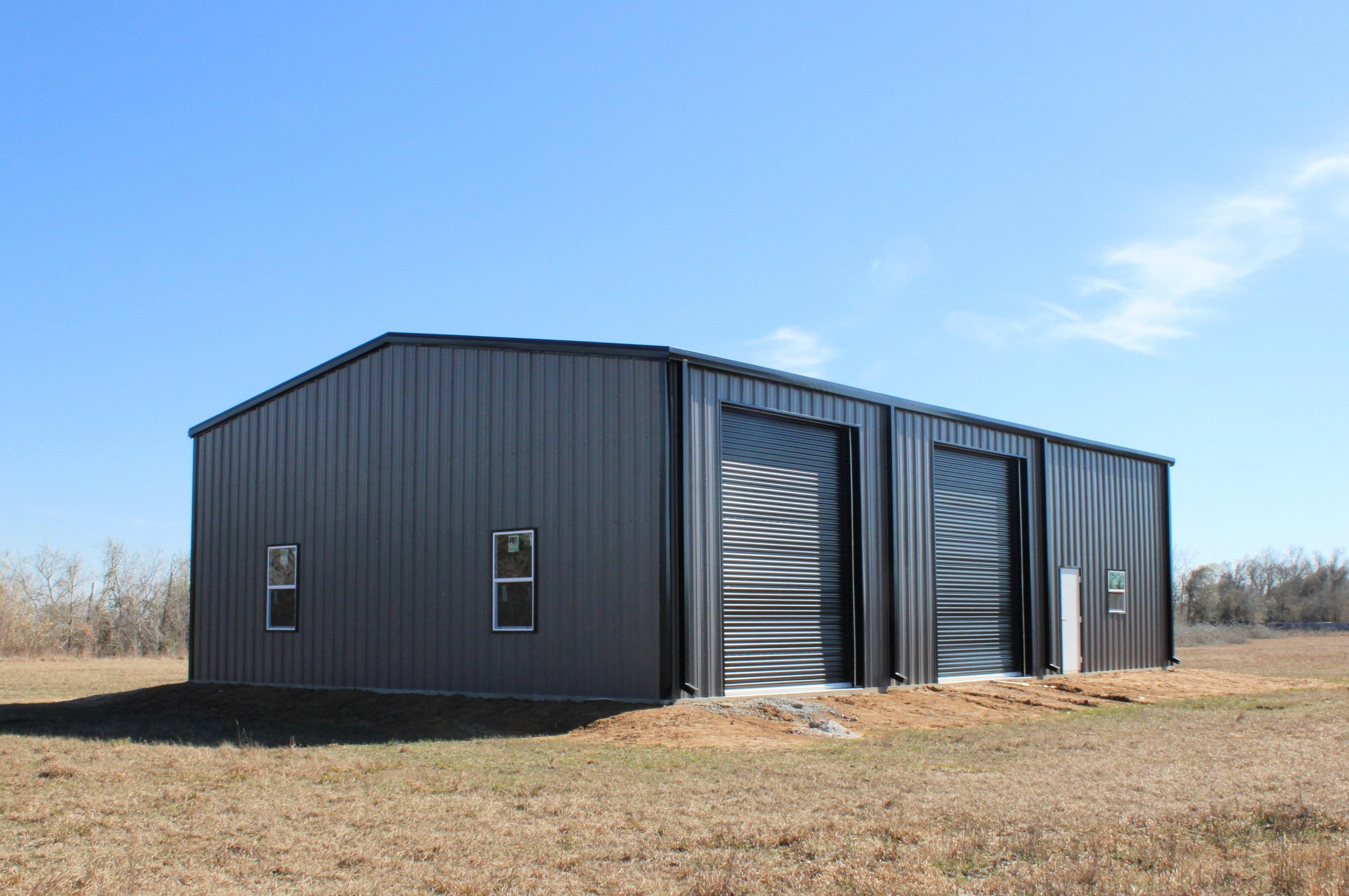 Angled exterior view of a custom 60’ x 40’ fully insulated metal shop in Wallis, TX featuring multiple black insulated roll-up doors, residential windows, and a durable Texas-ready design.