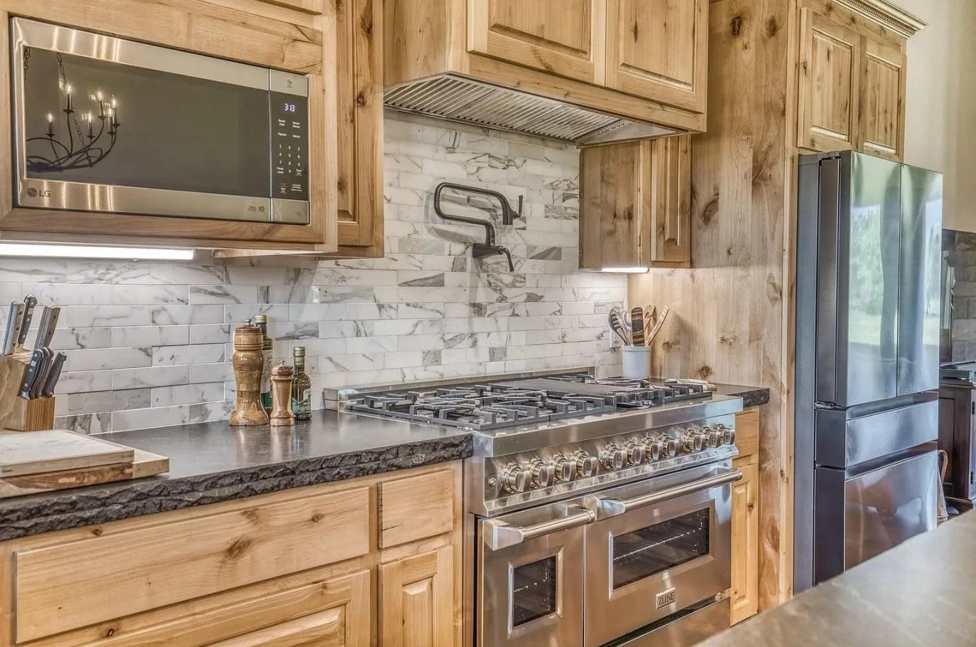Close-up of the kitchen’s professional-style gas range, stainless-steel appliances, natural wood cabinetry, stone countertops, and marble-look tile backsplash.