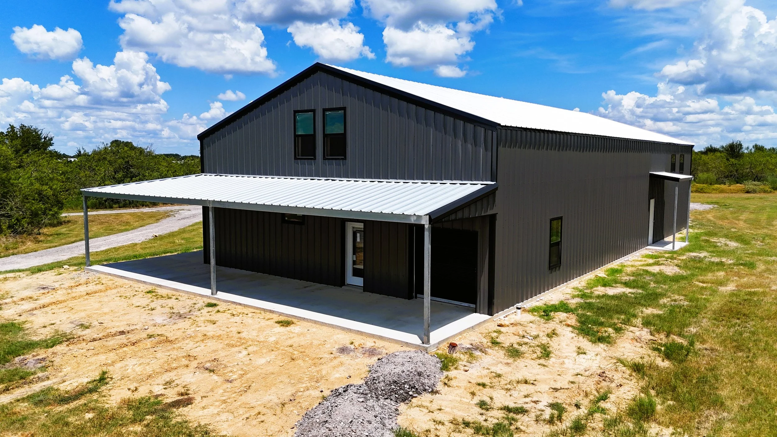 Aerial front perspective of a custom weld-up shop and home barndominium featuring an expansive L-shaped porch, Galvalume roof, and charcoal gray metal exterior in Burton, TX.