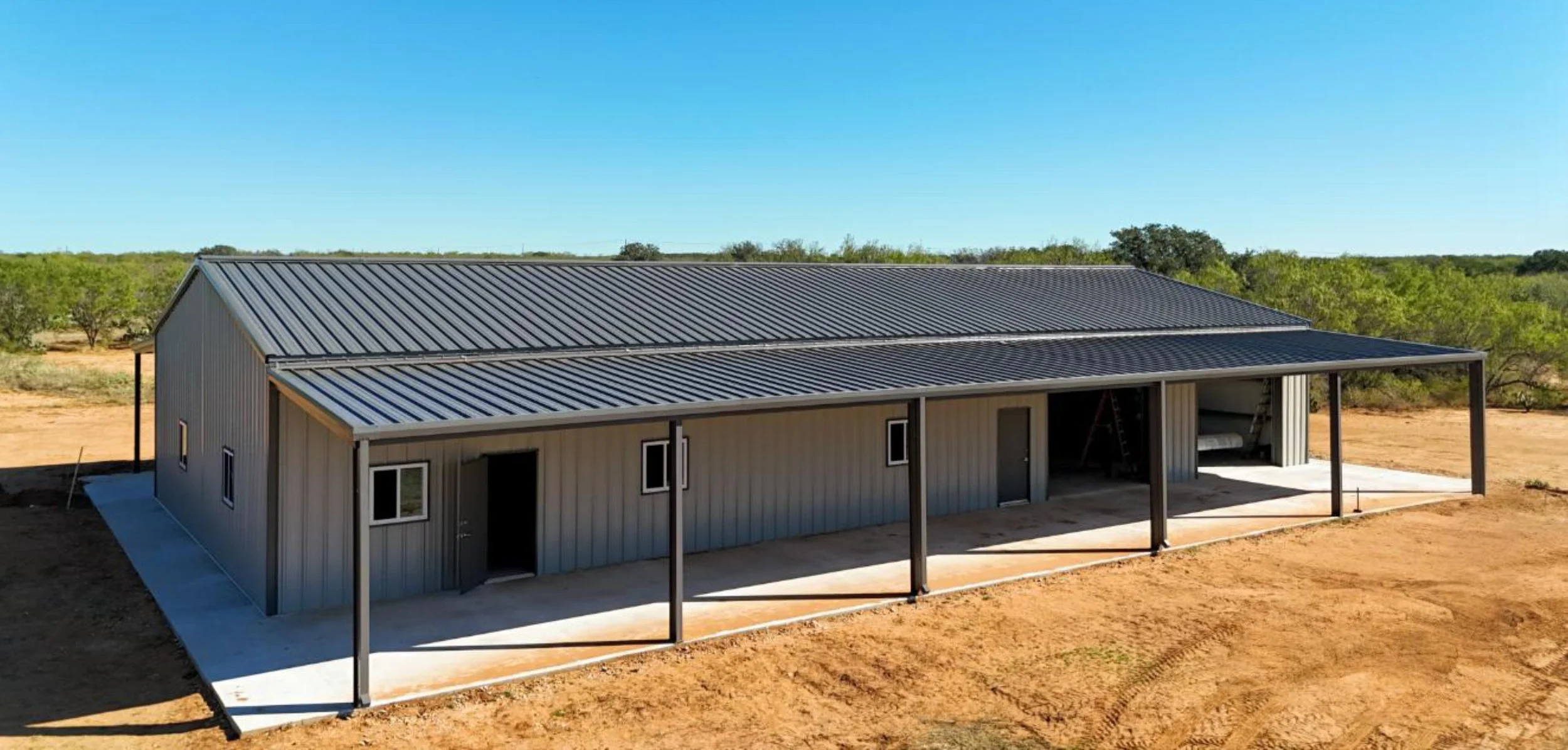 Covered lean-to porch space on metal barndominium with walk doors and windows, showing shade structure supported by steel posts.
