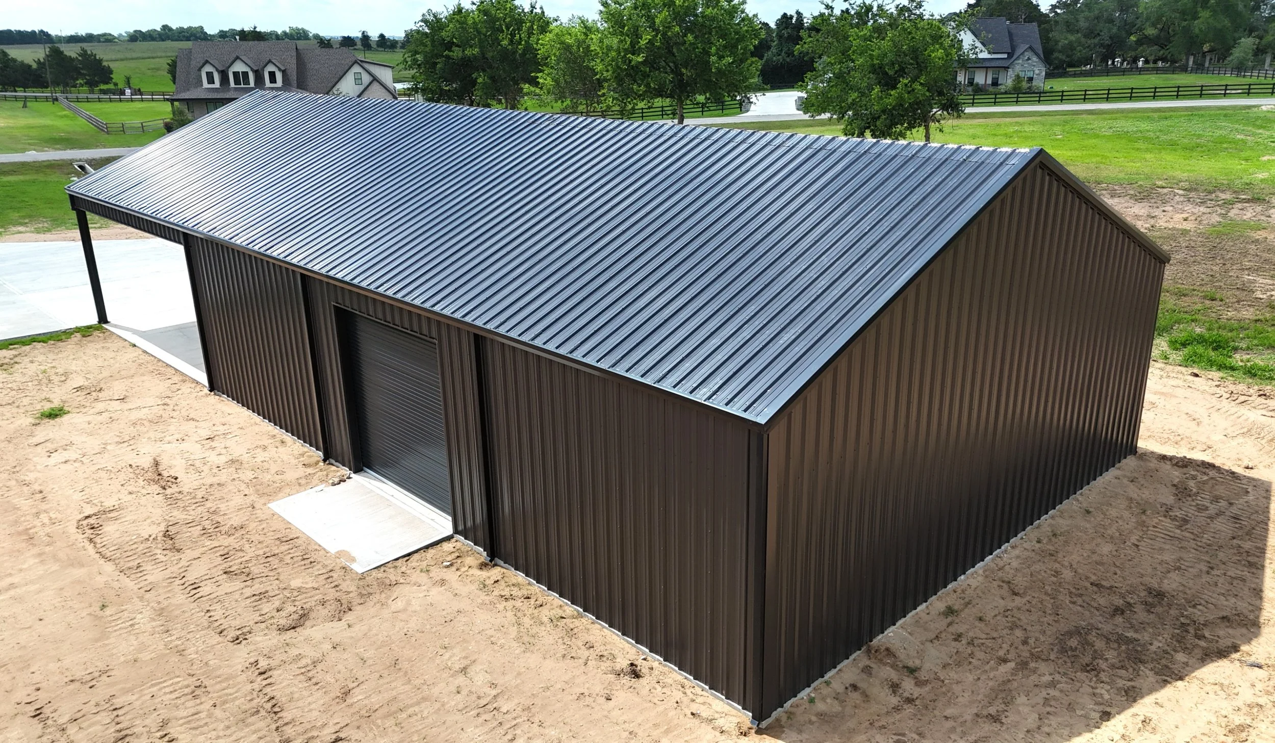 Aerial side view of custom 60x40x14 metal shop in Chappell Hill, Texas featuring Burnished Slate steel siding, black R-panel roof, steep 5/12 roof pitch, and oversized roll-up door designed for equipment and vehicle access.
