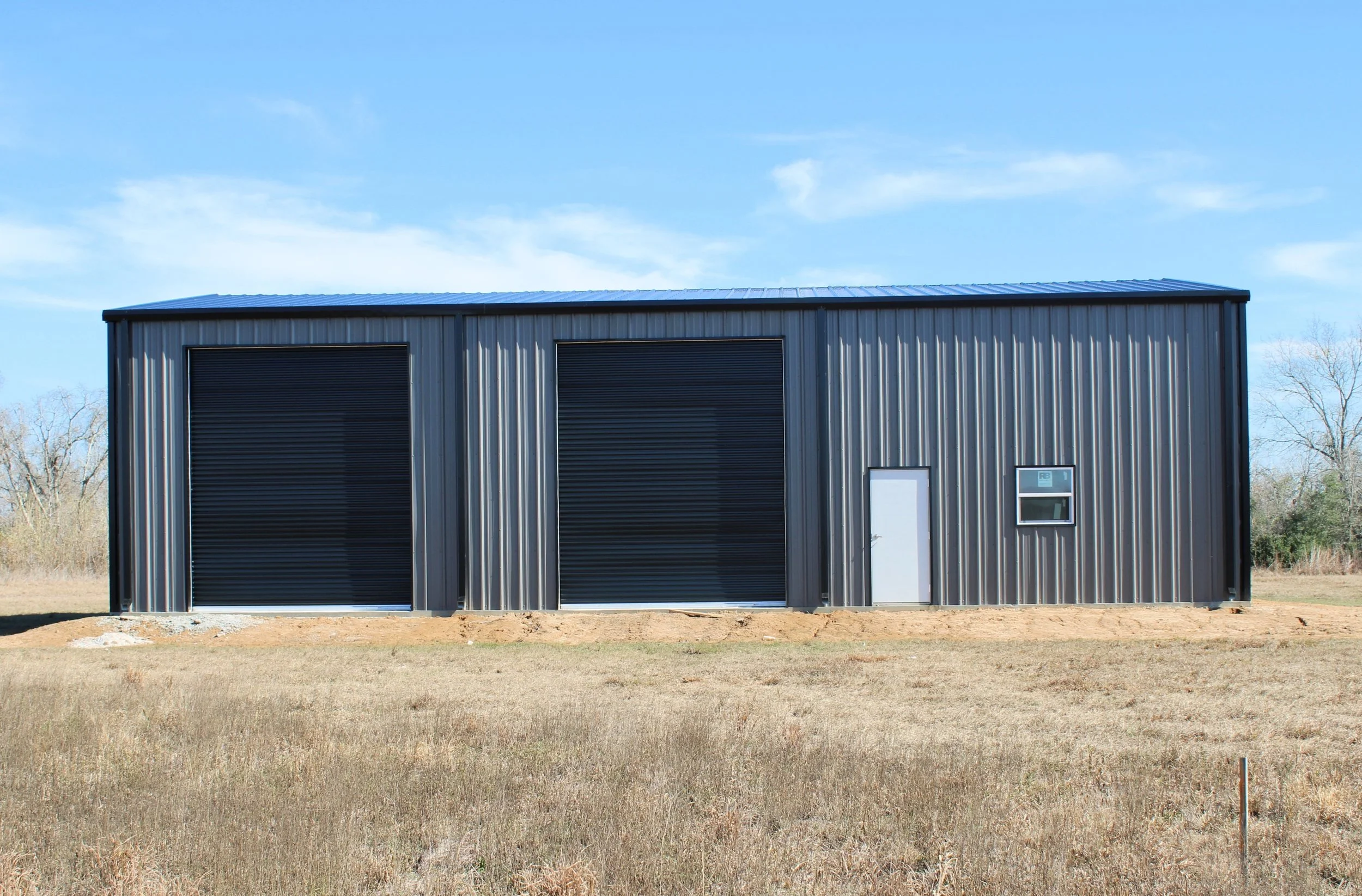 Front view of a custom 60’ x 40’ metal shop in Wallis, TX with three black insulated roll-up doors, white walk door, and clean modern exterior design.