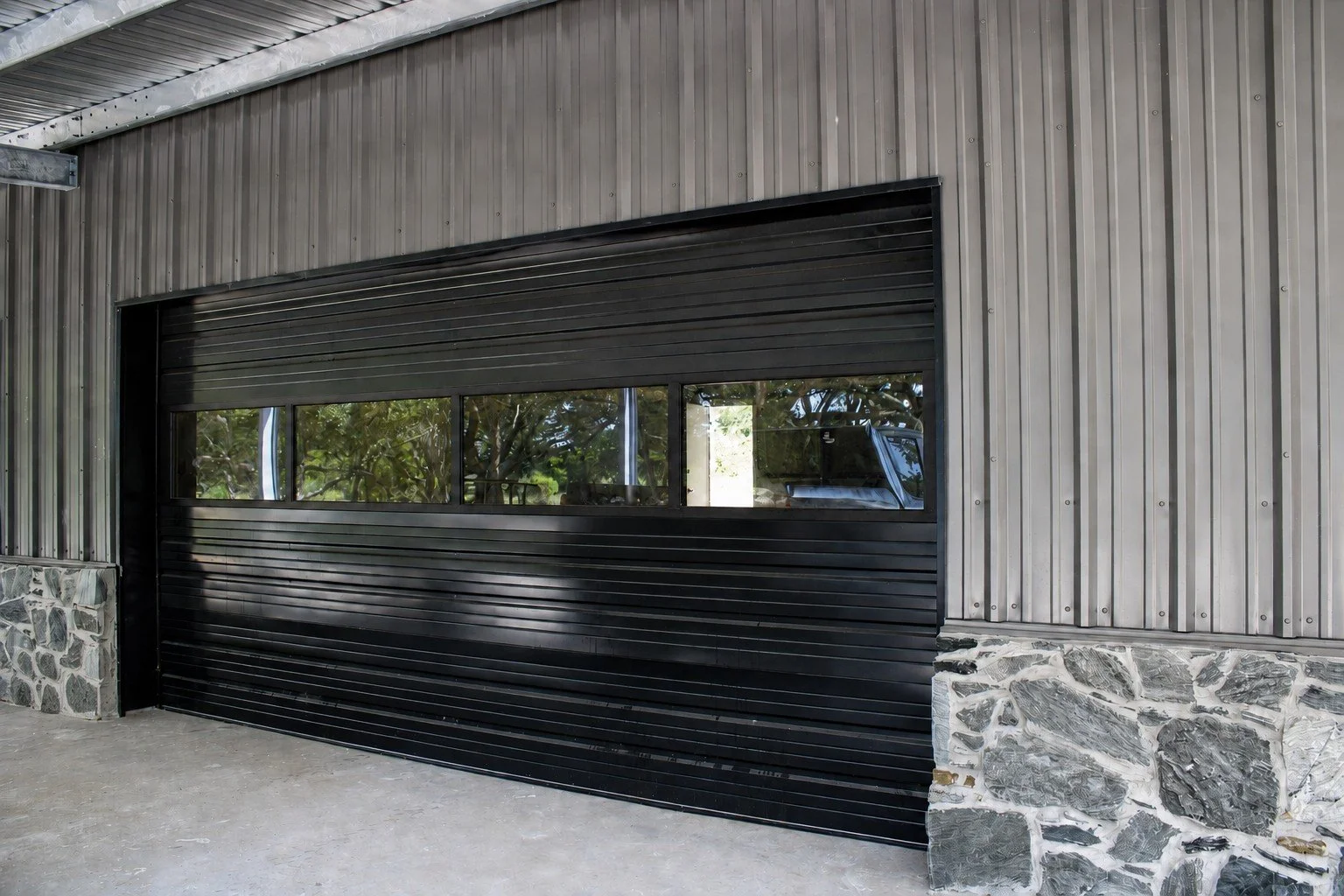 Black sectional garage door with glass panels, complementing the Charcoal Gray siding and black trim of this custom barndominium in Damon, Texas.