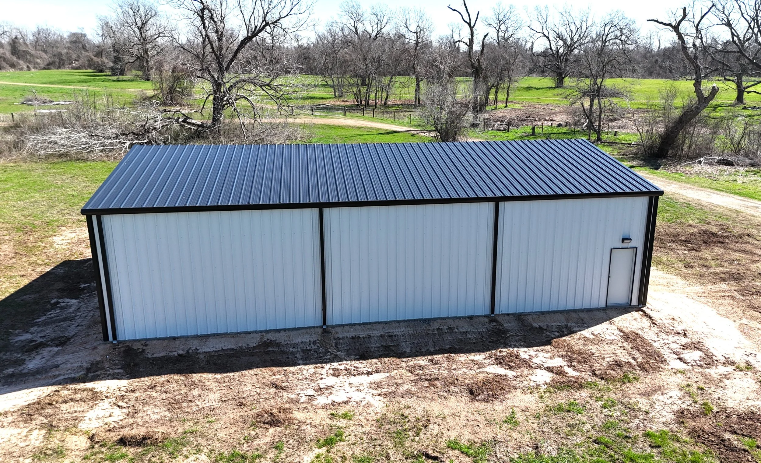Aerial view of 2/12 pitch black metal roof on custom shop in Fulshear, Texas with full gutter and downspout system installed for drainage control, and walk door.