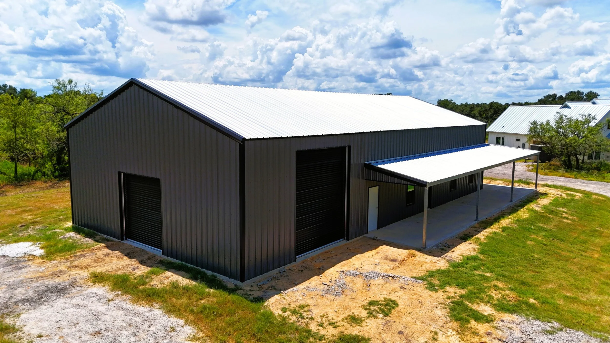 Rear angle of a custom 40x80 weld-up shop and home barndominium with insulated black roll-up doors, charcoal gray siding, and an L-shaped covered porch in Burton, TX.