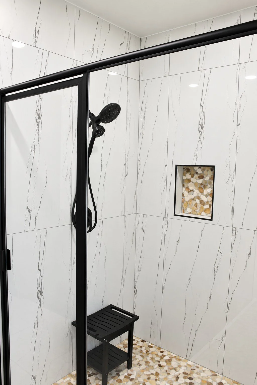 Large tiled walk-in shower with a black-framed glass enclosure, handheld shower fixture, built-in niche, and pebble tile floor.