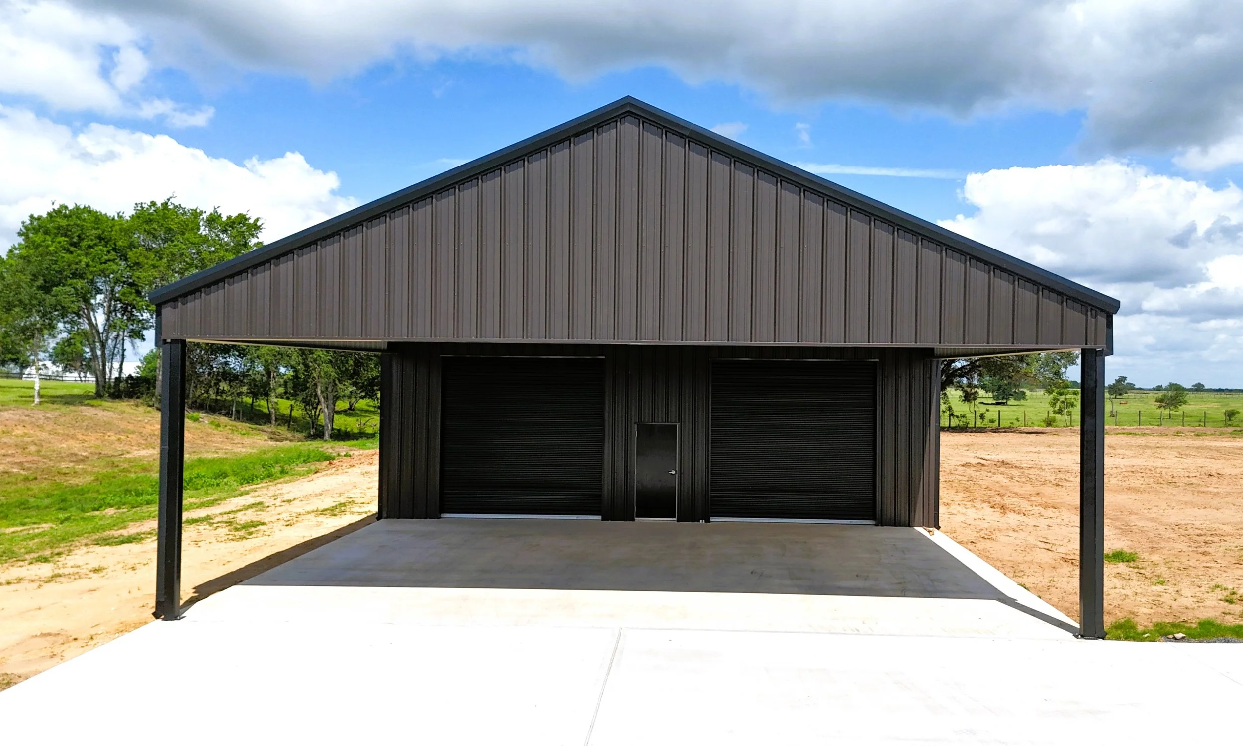 Front view of custom 60x40x14 metal shop in Chappell Hill, Texas featuring a 5/12 roof pitch, oversized 30x40 covered awning, black roll-up doors, Burnished Slate siding, and black trim on reinforced concrete slab.