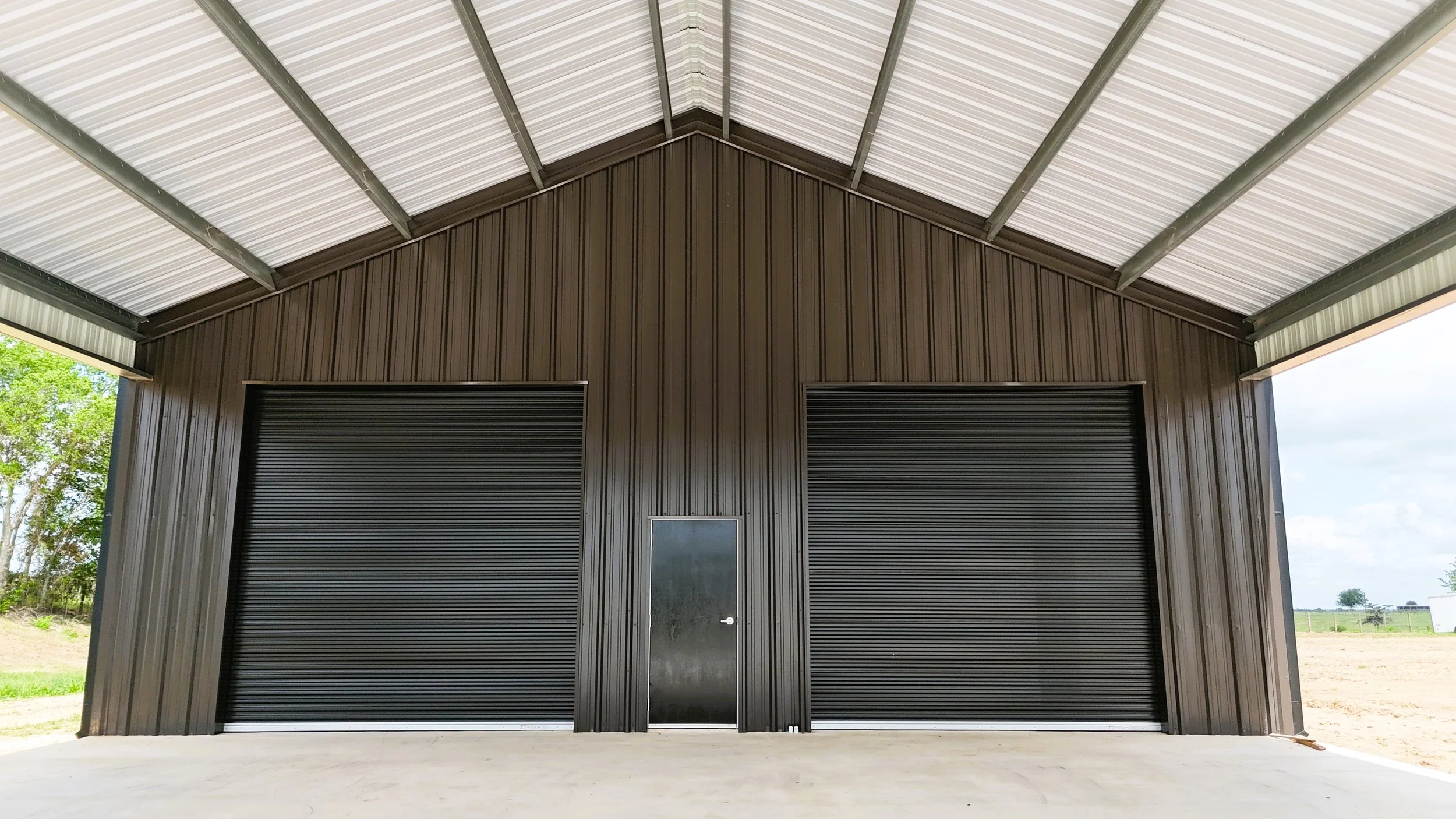 Angled aerial view of custom insulated metal shop with oversized awning and black roof in Washington County, Texas built for equipment storage, workshop space, and outdoor entertaining.