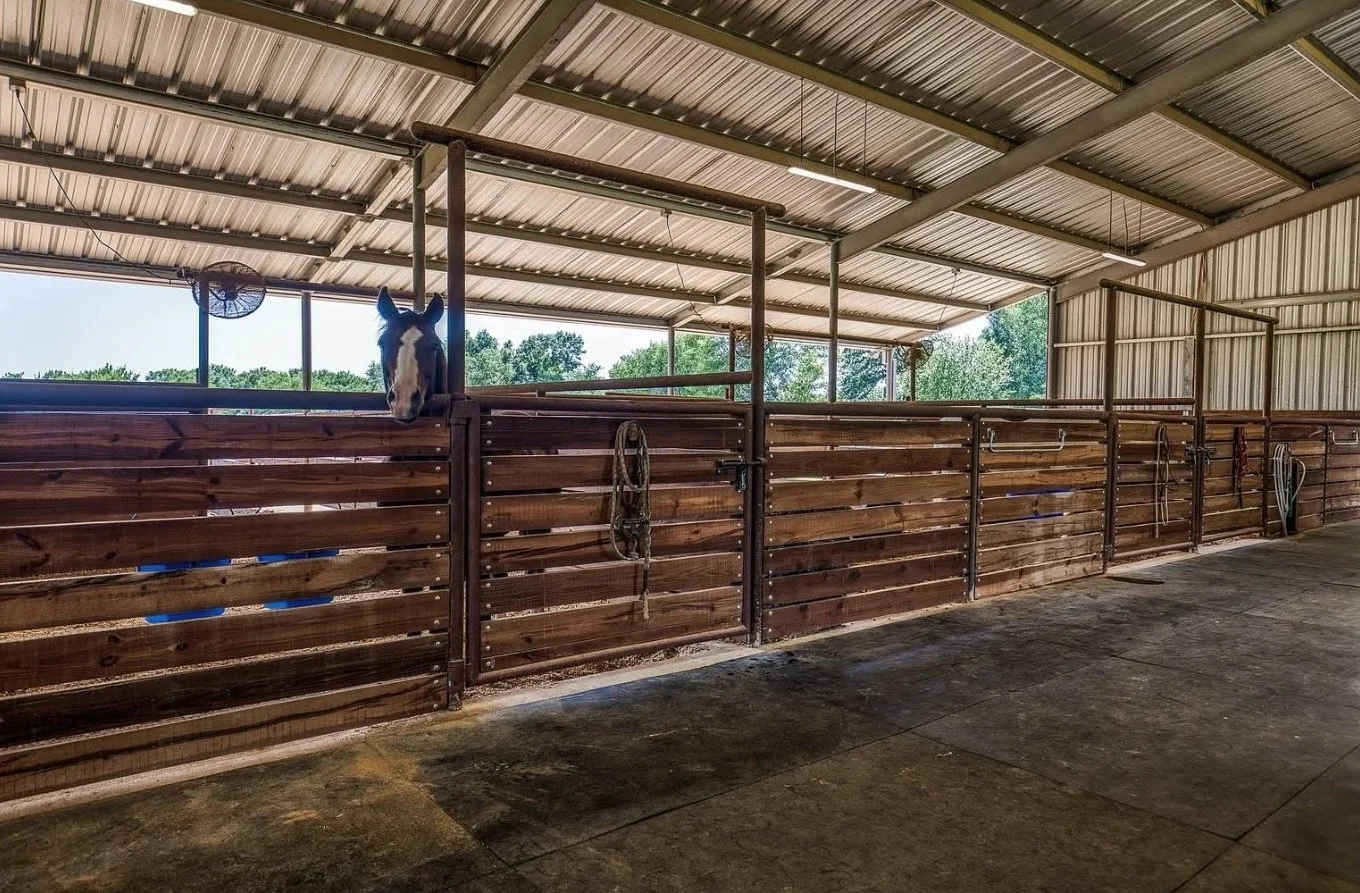 Custom covered horse stalls constructed with steel framing and wood boards, providing shade, ventilation, and convenient access from the barn aisle.