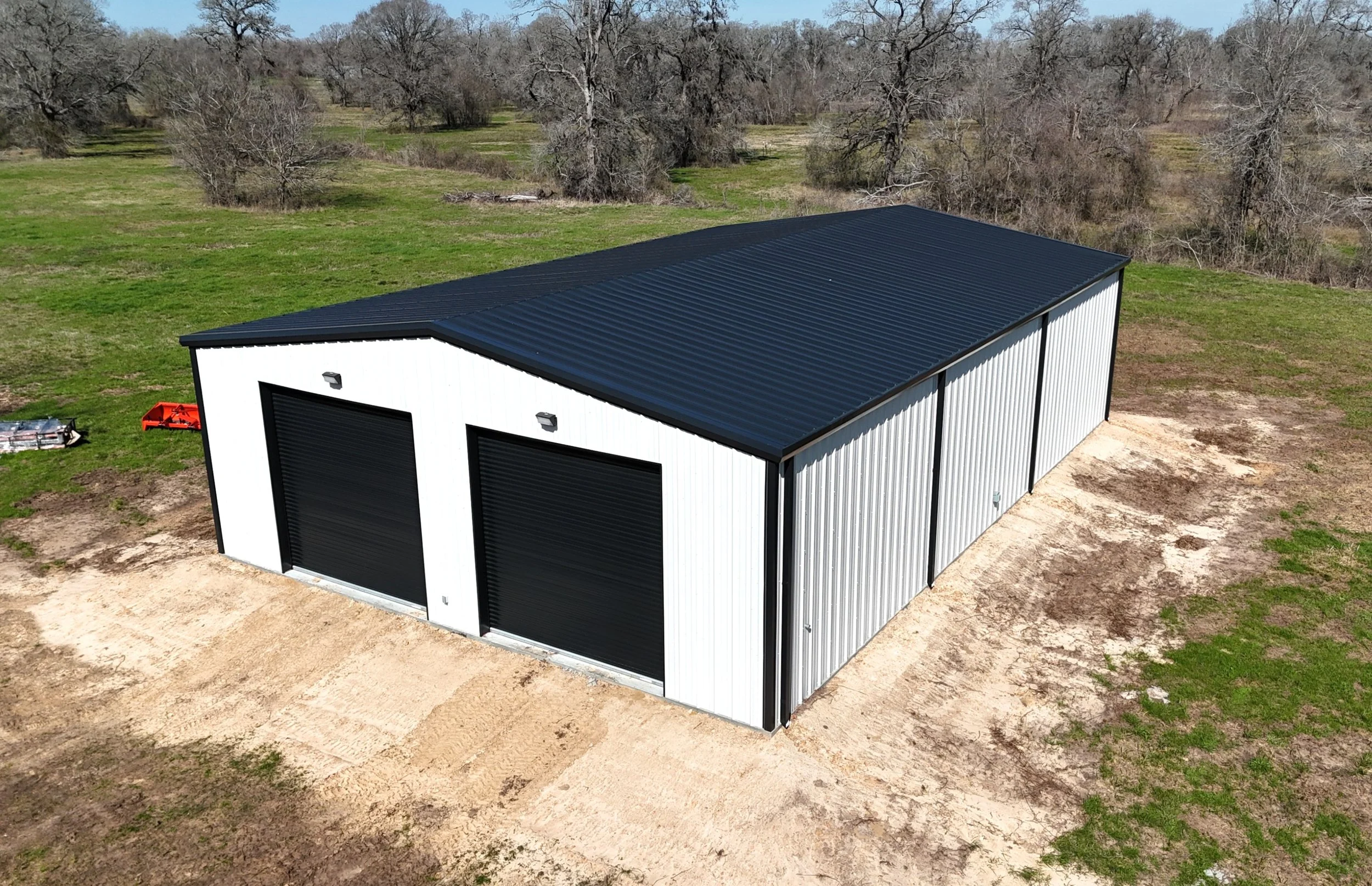 Front aerial view of custom metal workshop in Fulshear, TX featuring dual 14’ x 14’ black roll-up doors, walk-through entry, and classic black-on-white finish.