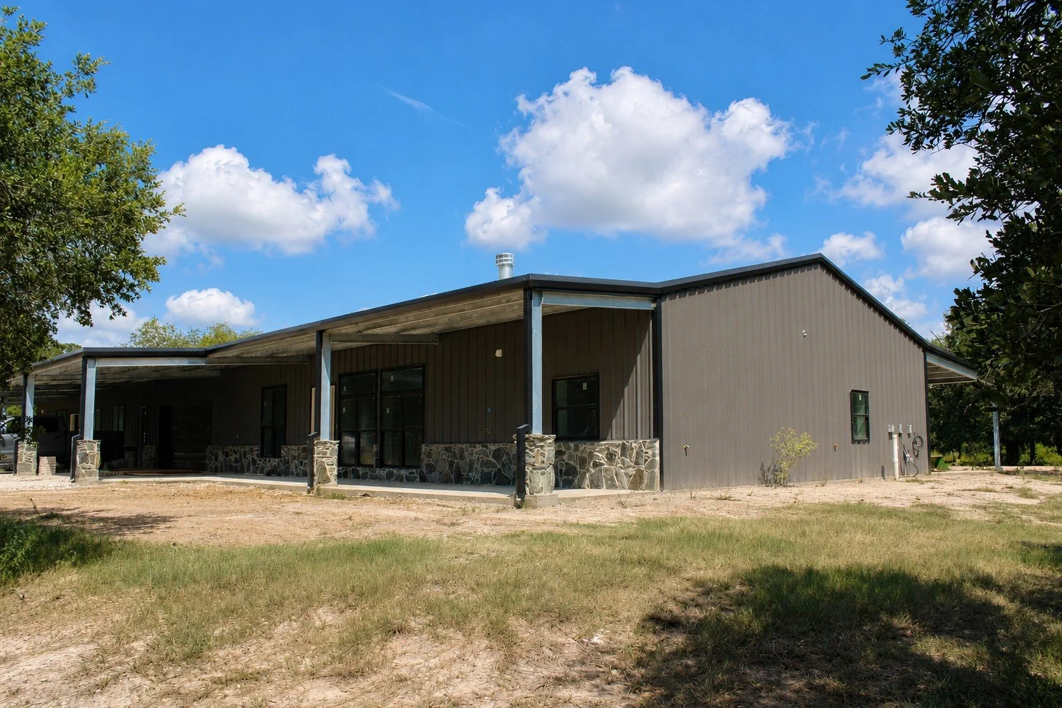 Wide exterior view of the dual-residence barndominium featuring Charcoal Gray metal siding, black trim, covered porches, and custom stonework.