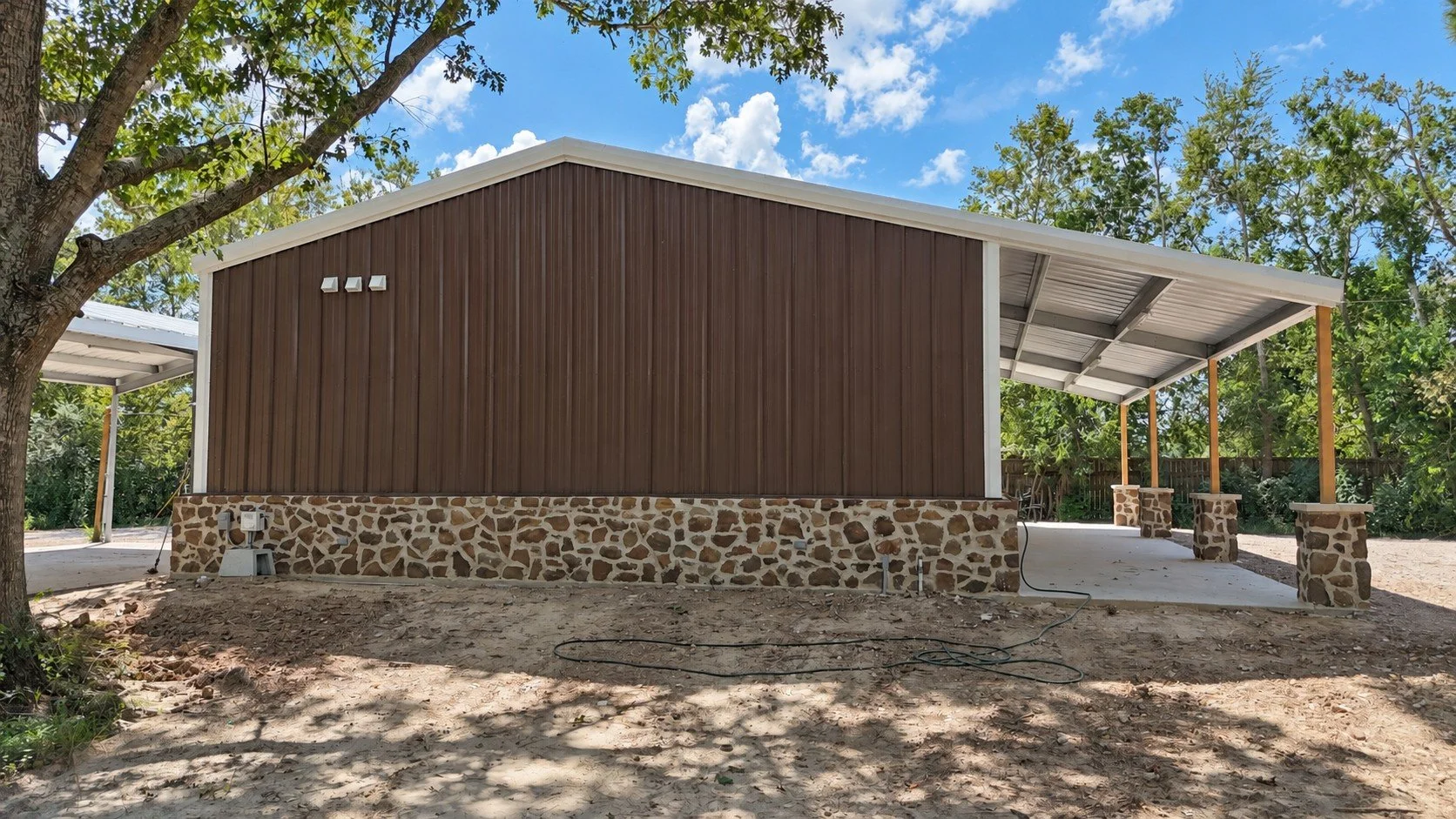 Side exterior of a windstorm-certified barndominium with brown metal siding, stone wainscoting, and a large covered concrete patio in Damon, Texas.