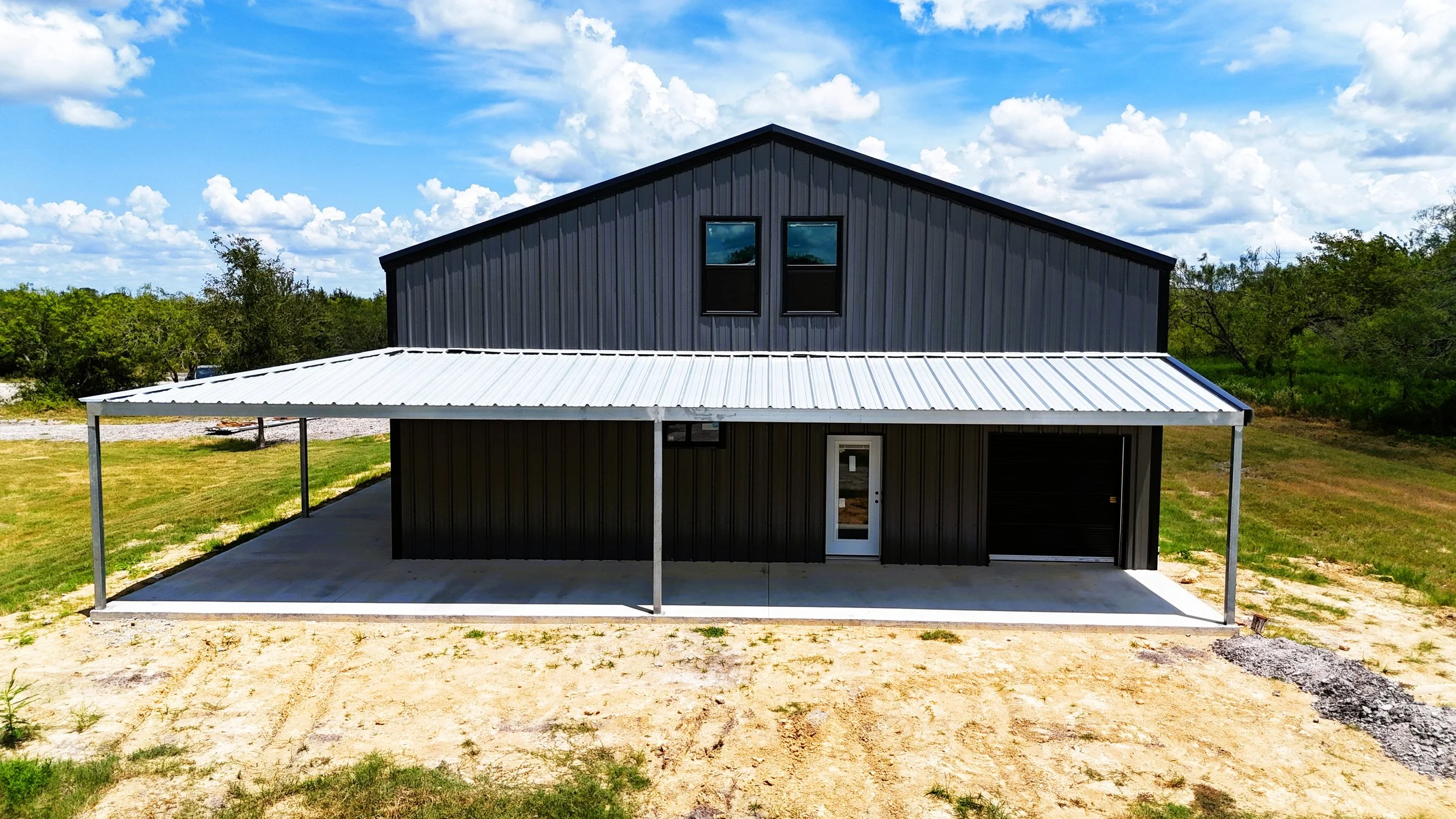 Front elevation of a custom steel barndominium showing the covered porch, black-framed windows, insulated roll-up door, and modern charcoal gray exterior.