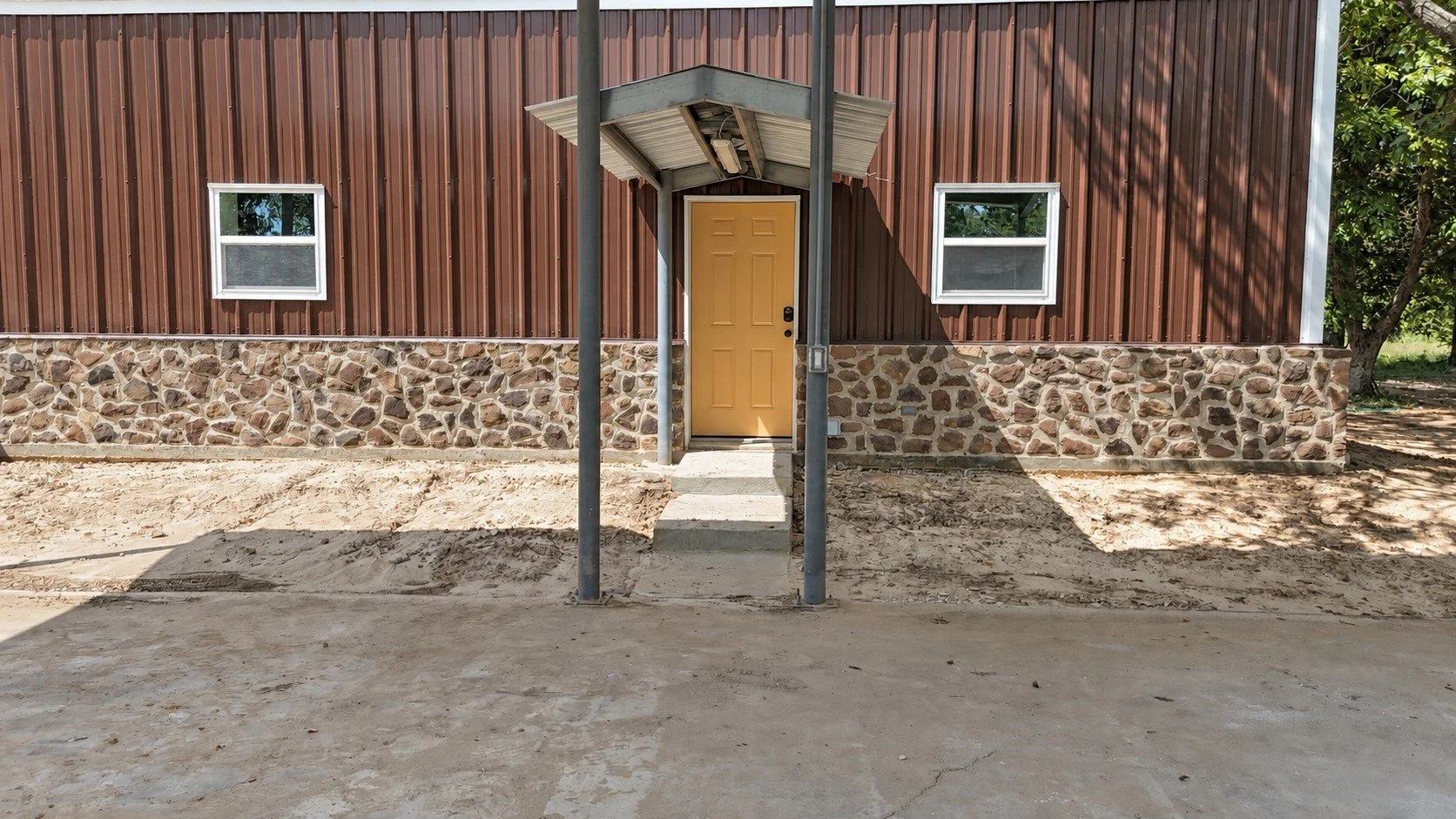Covered rear entrance of the Damon barndominium featuring a mustard-colored door, metal entry awning, stone exterior, and concrete walkway.