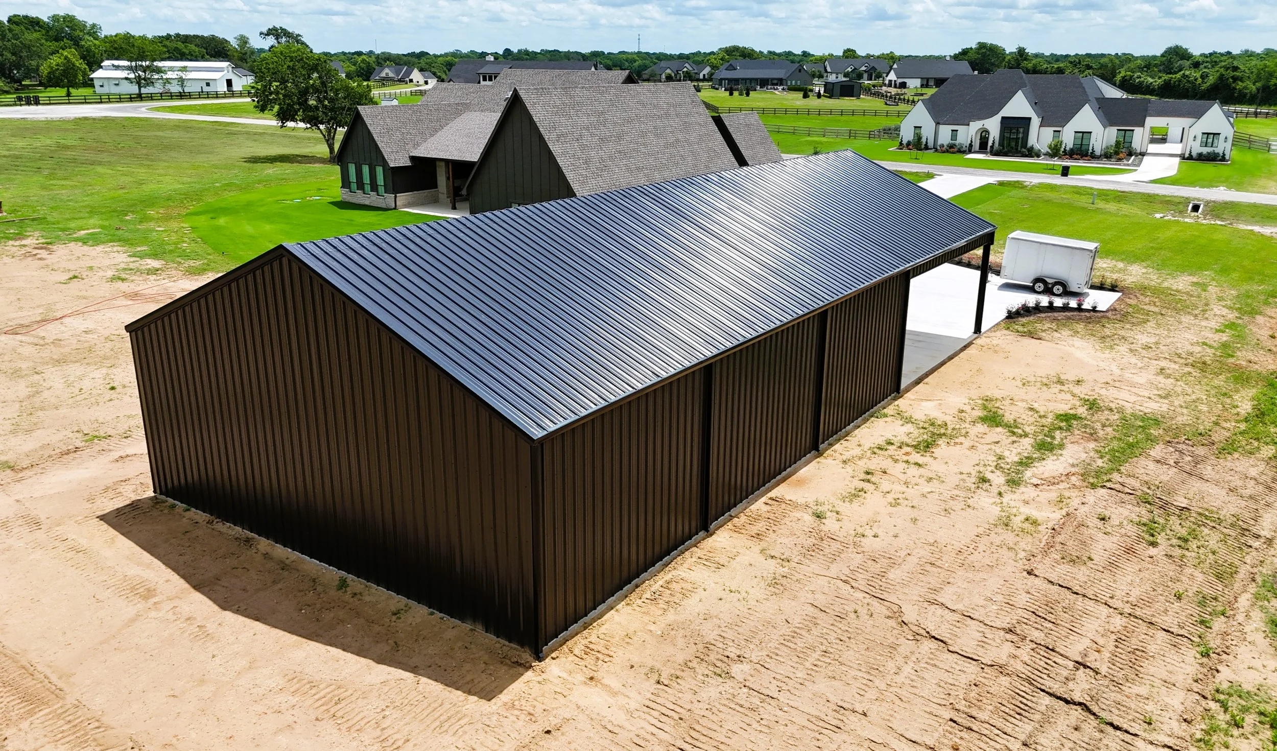 Aerial view of custom weld-up metal building with Burnished Slate siding and black roof in Chappell Hill, TX featuring a large covered awning, modern residential-style roof pitch, and spacious concrete driveway.