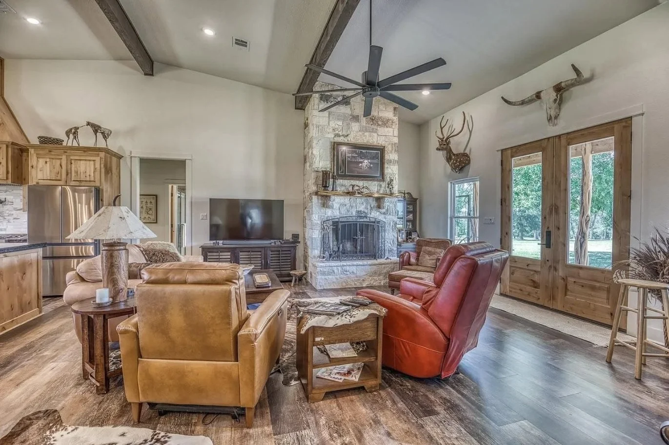 Vaulted barndominium living room featuring exposed wood beams, a floor-to-ceiling stone fireplace, oversized ceiling fan, and double doors leading outside.