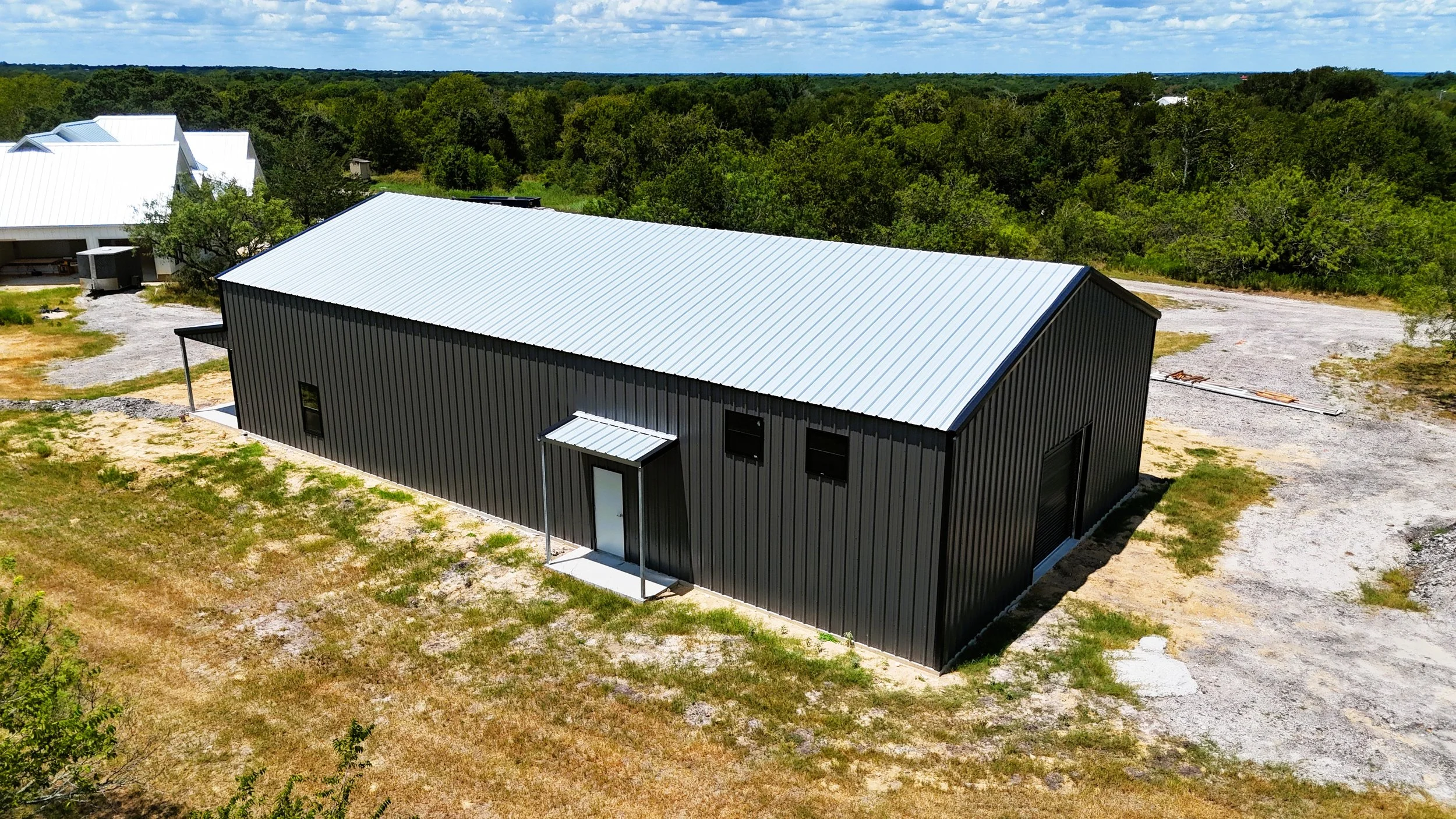 Aerial rear view of a custom 40x80 two-story barndominium with Galvalume roof, charcoal gray metal siding, black trim, and covered rear entry in Burton, Texas.