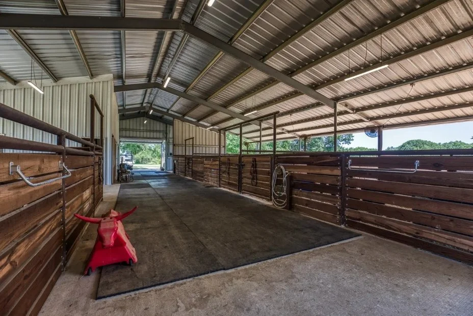 Interior view of the four-stall horse barn featuring a wide concrete aisle, custom stalls, overhead lighting, fans, and convenient wash and tie areas.