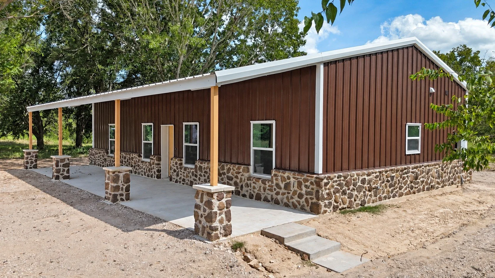 Angled exterior view of the Damon, Texas barndominium featuring brown metal siding, white trim, custom stonework, covered porches, and natural wood posts.