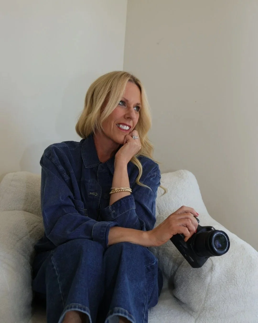A blonde woman sitting on a white couch, smiling and looking to the side, holding a camera in her right hand and resting her chin on her left hand, wearing a denim jacket and jewelry.