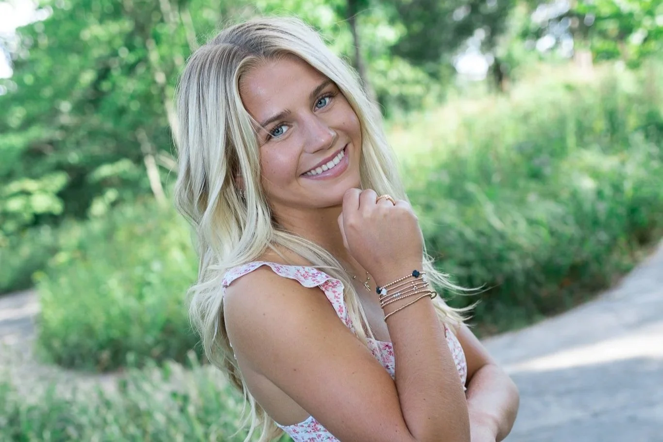 A young woman with long blonde hair smiling outdoors in a natural setting with greenery.