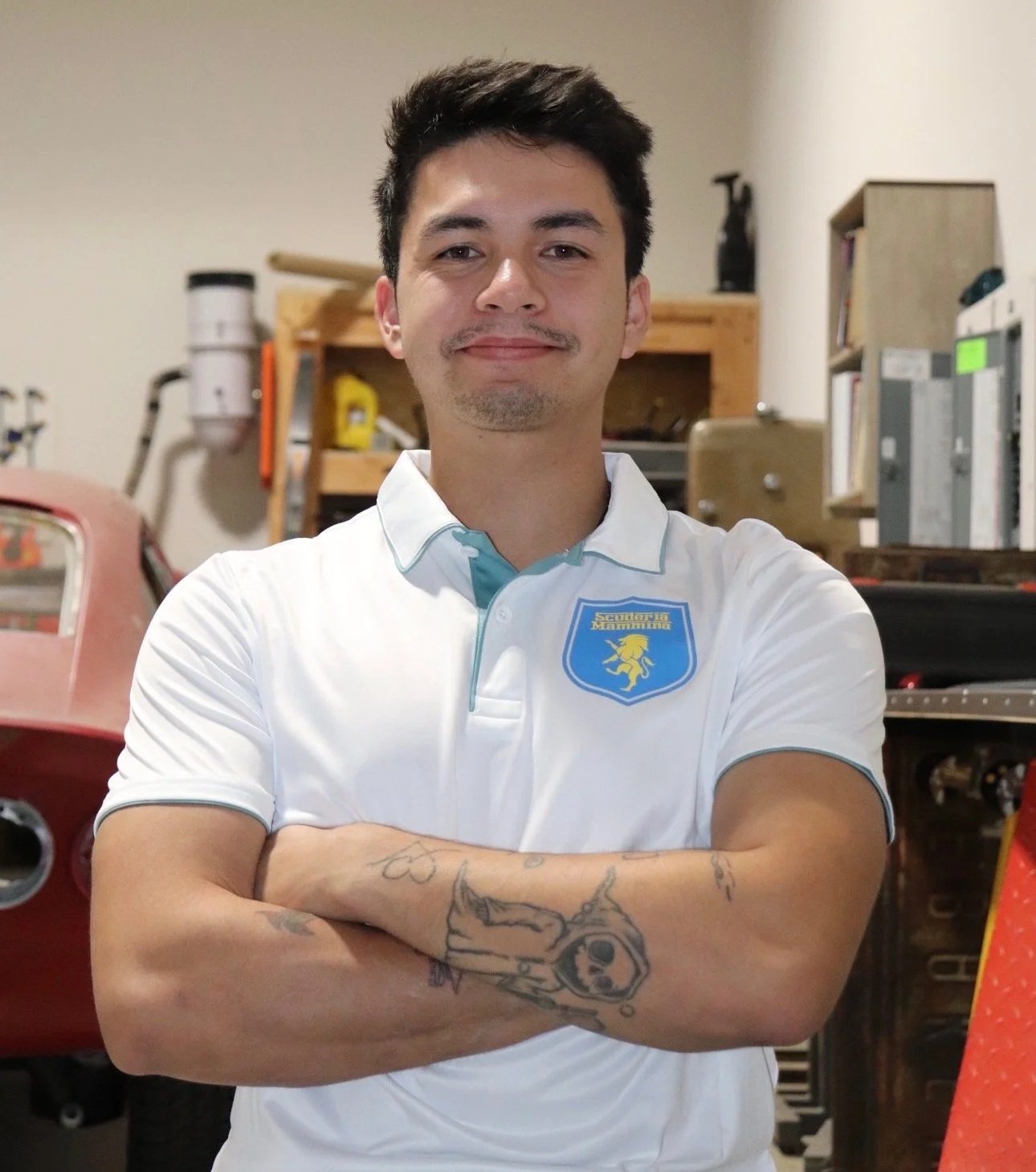 A man with dark, curly hair, gray glasses, and a beard and mustache, wearing a dark green sweater and a white shirt underneath, sitting indoors with neutral background.