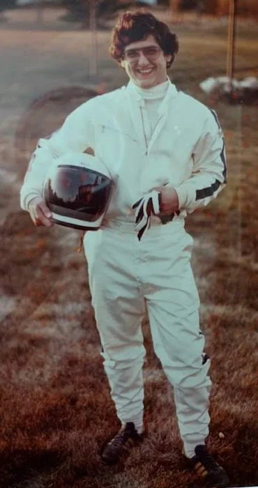 A young person dressed in a white racing suit holding a black helmet, standing outdoors on a grassy field with a smile.
