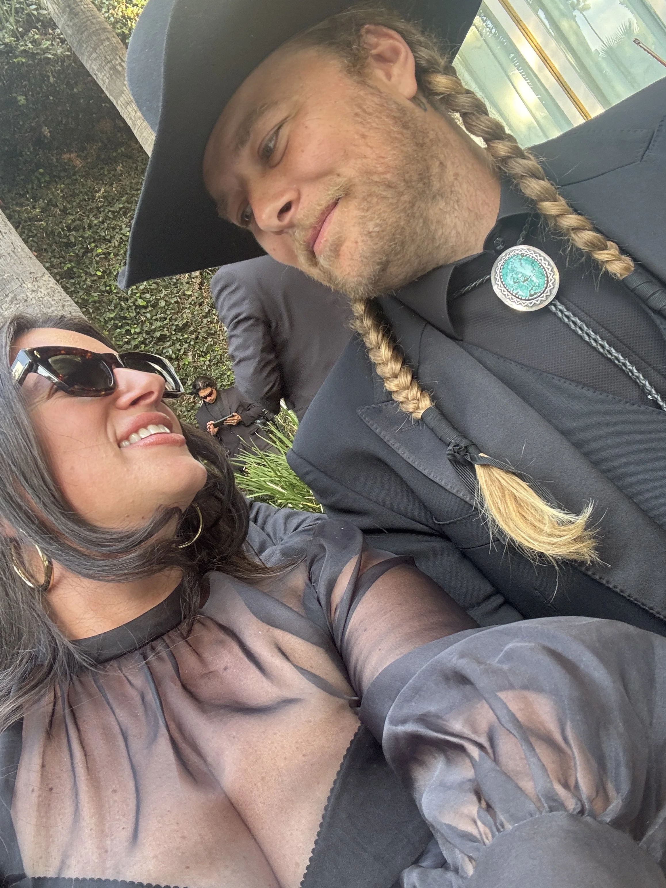 A woman with dark hair and sunglasses smiling, taking a selfie with a man dressed as a cowboy in a black hat, black shirt with a bolo tie, and a braid, at an outdoor event.