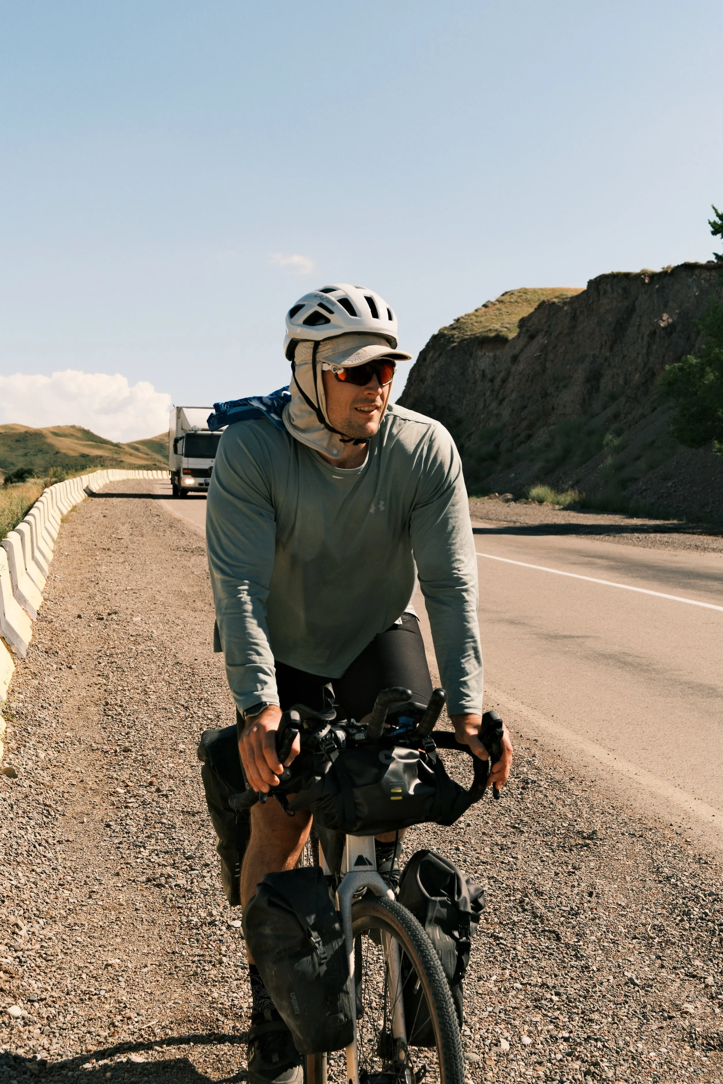 A man wearing a helmet and sunglasses riding a bicycle along a road with a guardrail, hills, and a truck in the background on a sunny day.