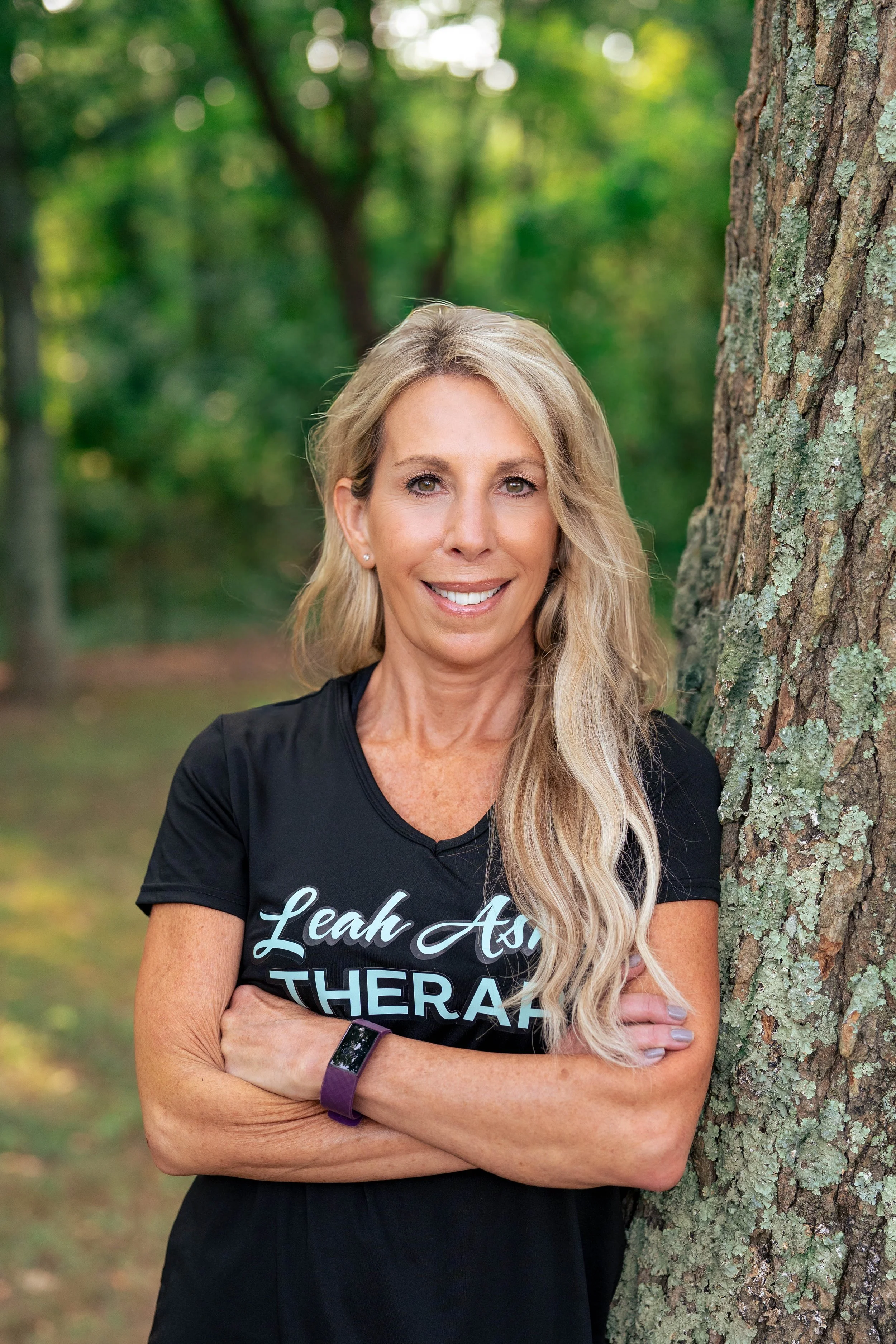 Portrait of Jessica Caldwell. A smiling woman with long blonde hair standing outdoors next to a tree in a forest setting, crossing her arms.