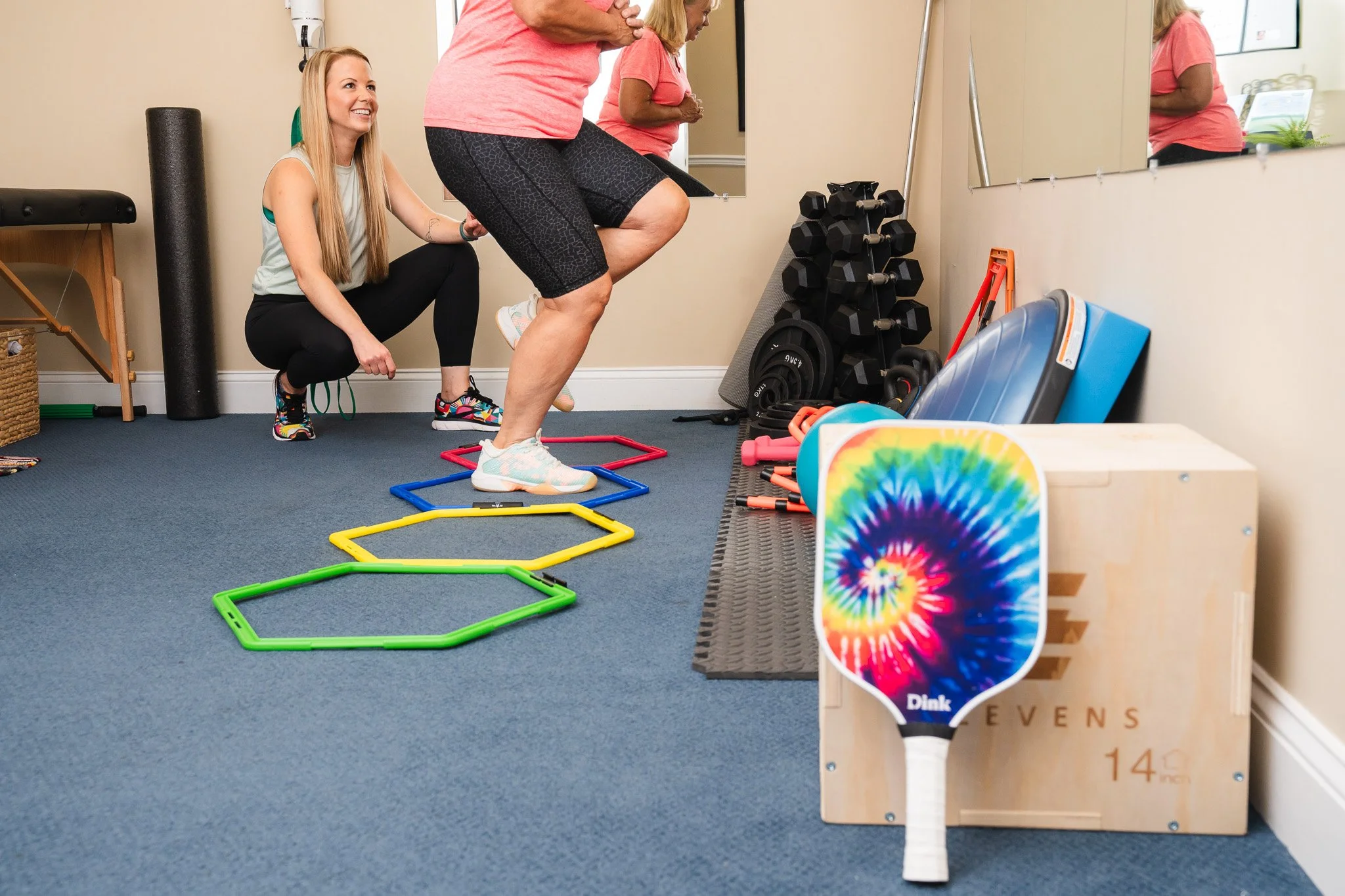 A woman in workout clothes is doing a step exercise on colorful individual steps in a fitness room. An instructor or trainer is kneeling beside her, providing guidance.