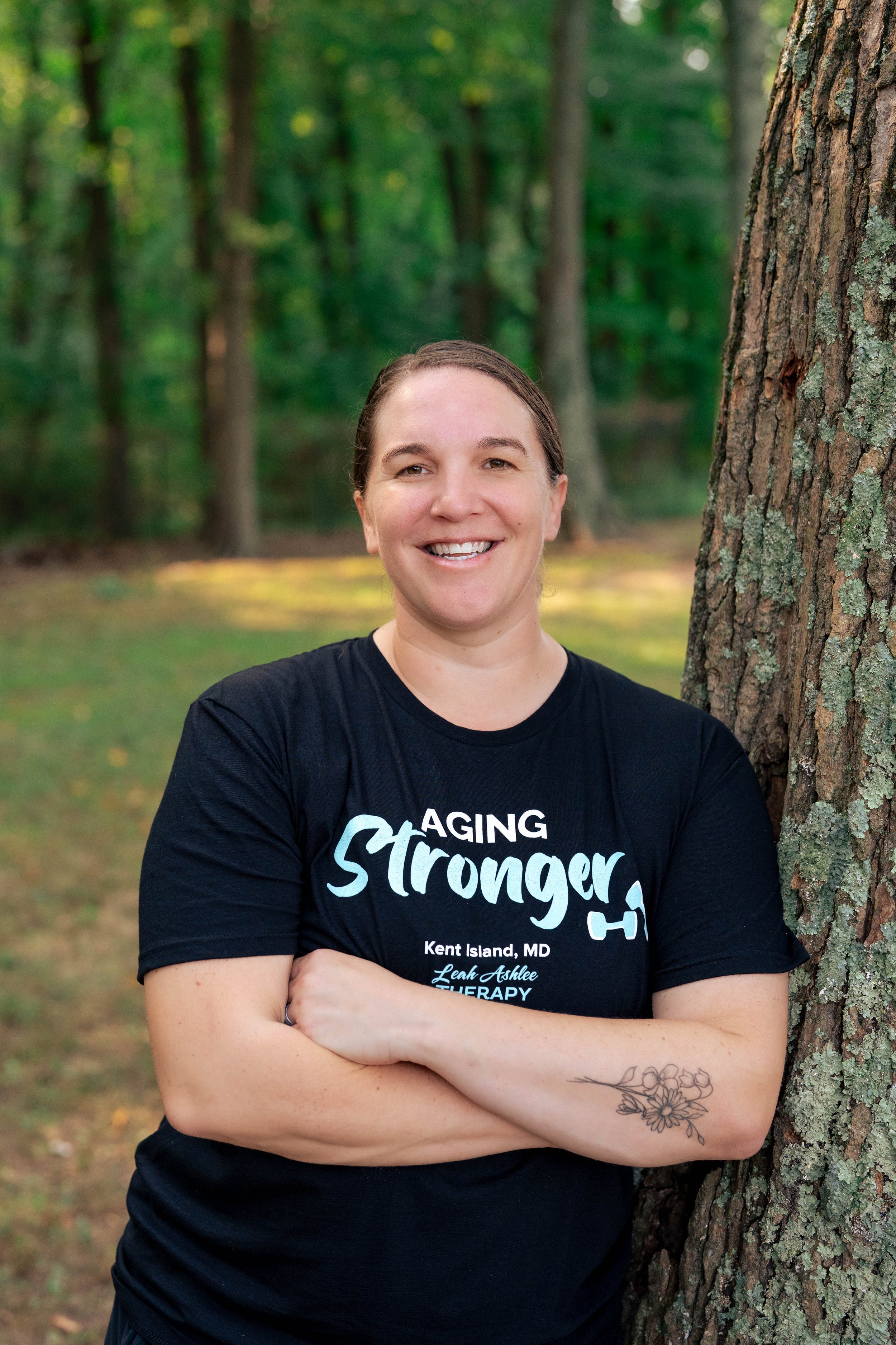 Portrait of Doctor Alyssa Fenn. A woman smiling with arms crossed stands beside a tree in a wooded area, wearing a black T-shirt that says 'Aging Stronger' and has additional text and a small graphic.