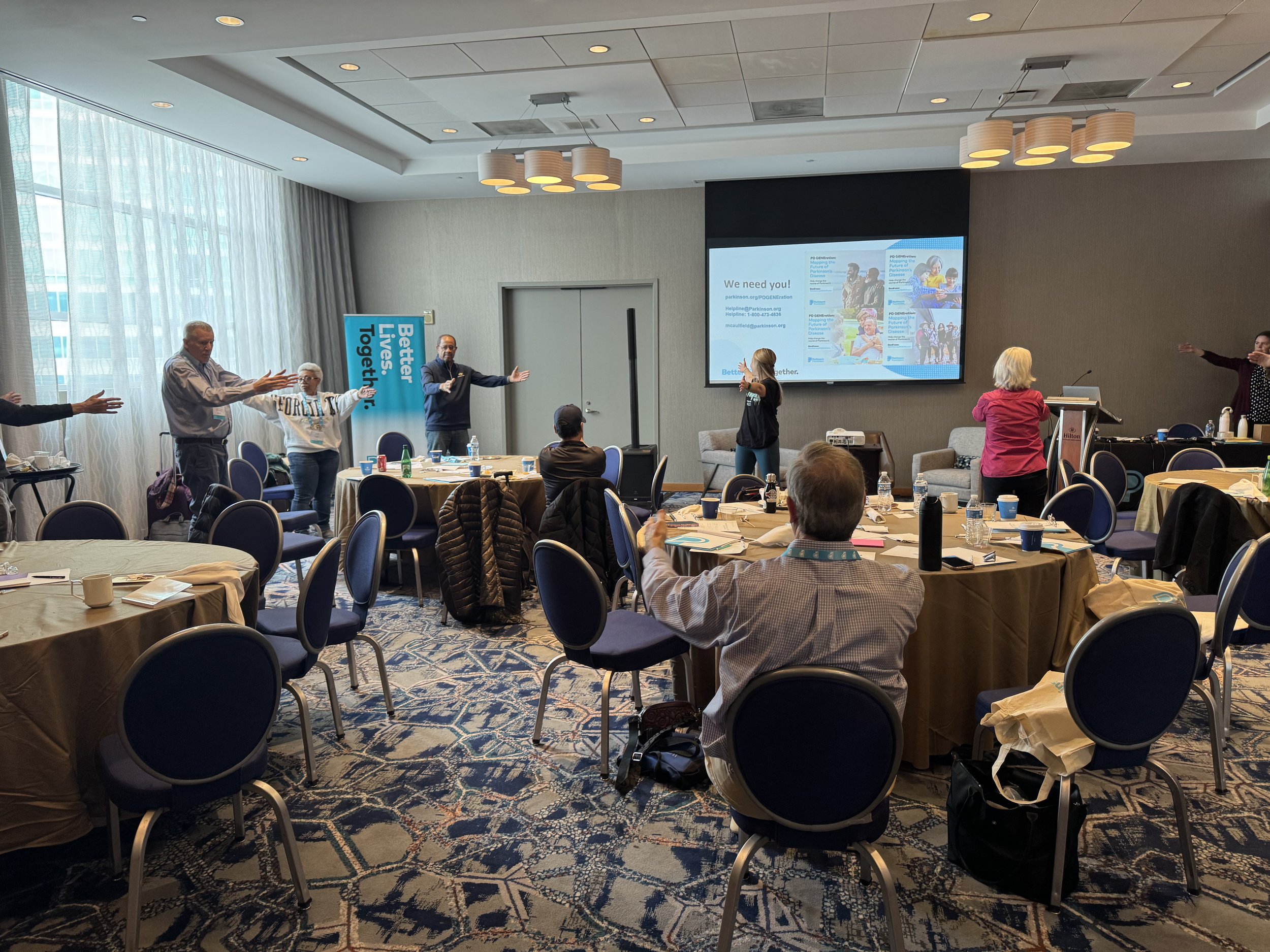 Group of people participating in a workshop or seminar in a conference room, with some standing at the front and others seated at round tables, while a presenter uses a large screen to display a presentation.