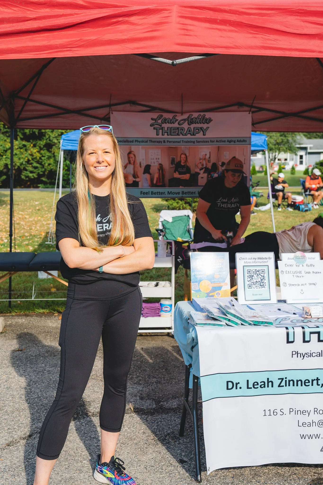 A woman standing in front of a booth promoting Leah Ashlee Therapy, smiling with crossed arms, wearing athletic clothes, at an outdoor event on a sunny day.