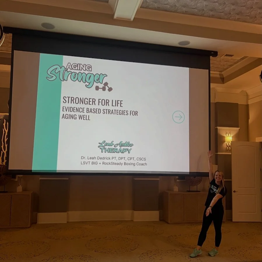 A woman standing in front of a large projector screen displaying a presentation titled 'Aging Stronger' with a subtitle 'Stronger for Life' and 'Evidence Based Strategies for Aging Well'. The presenter is smiling and pointing at the screen, and the room has a decorated ceiling with warm lighting.