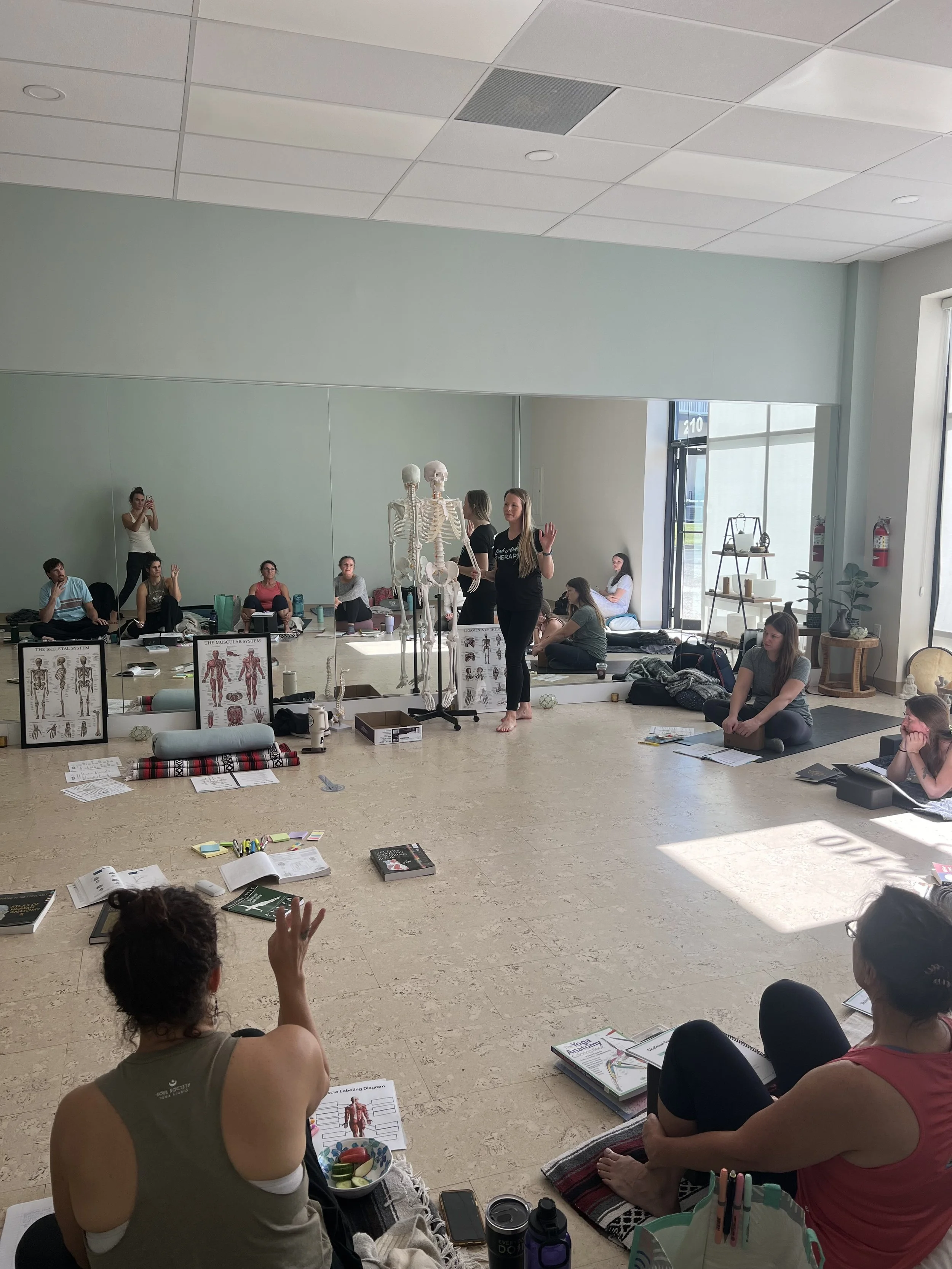A group of people participating in a workshop in a spacious room with large windows. A woman stands at the front, gesturing while speaking to the audience, with skeleton and anatomy charts displayed nearby. Participants sit on the floor with notebooks and materials around them.