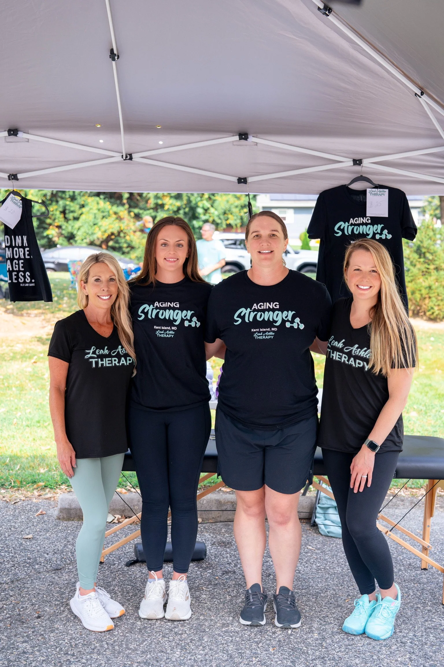 Four women standing together outside under a canopy, wearing black t-shirts with fitness or therapy-related messages. Two of the women have shirts reading 'Aging Stronger.' They are smiling, with trees and a few people in the background.
