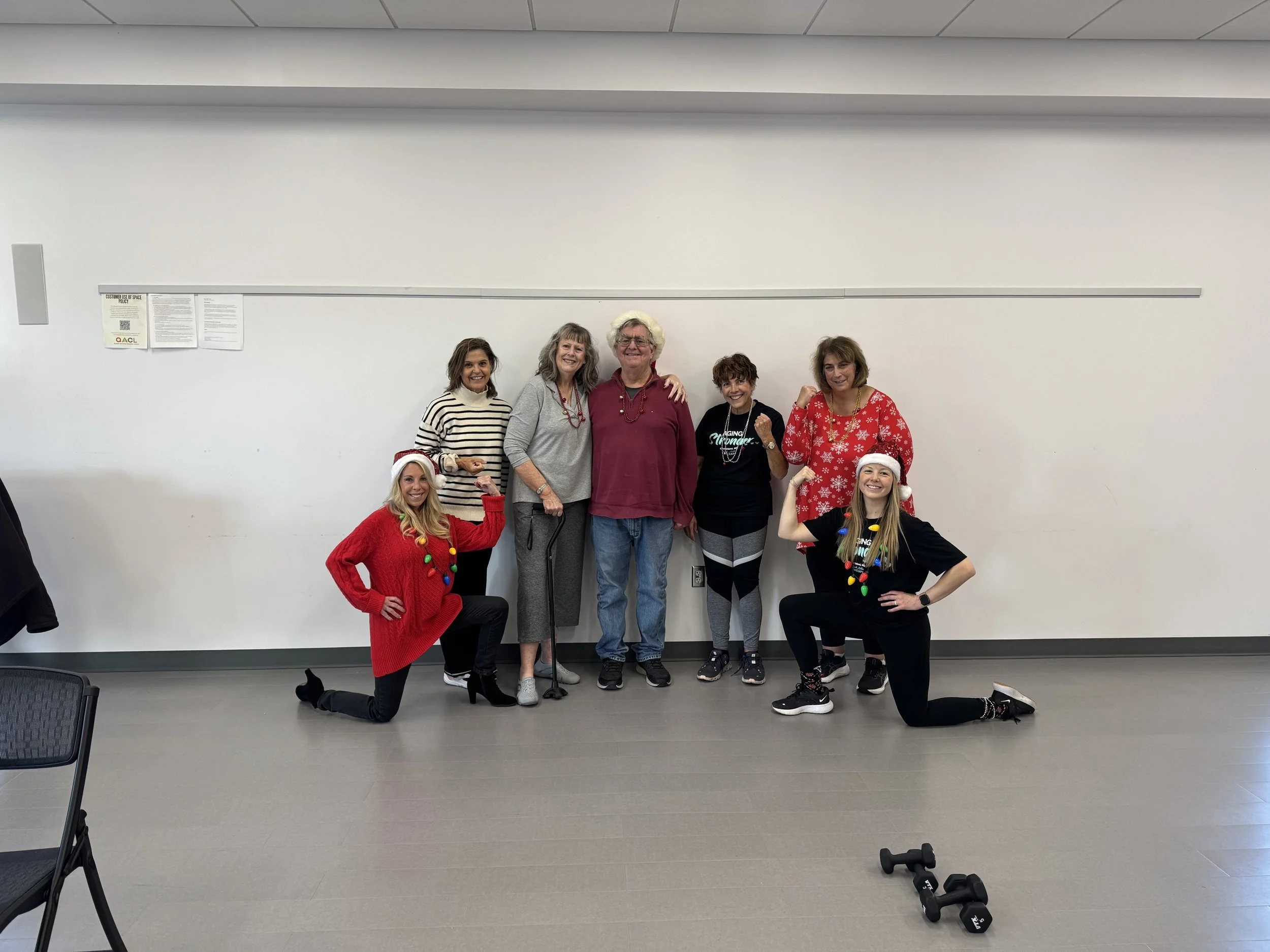 Group of men and women wearing Santa hats and festive sweaters, smiling and celebrating in a classroom setting with white walls and small black dumbbells on the floor.