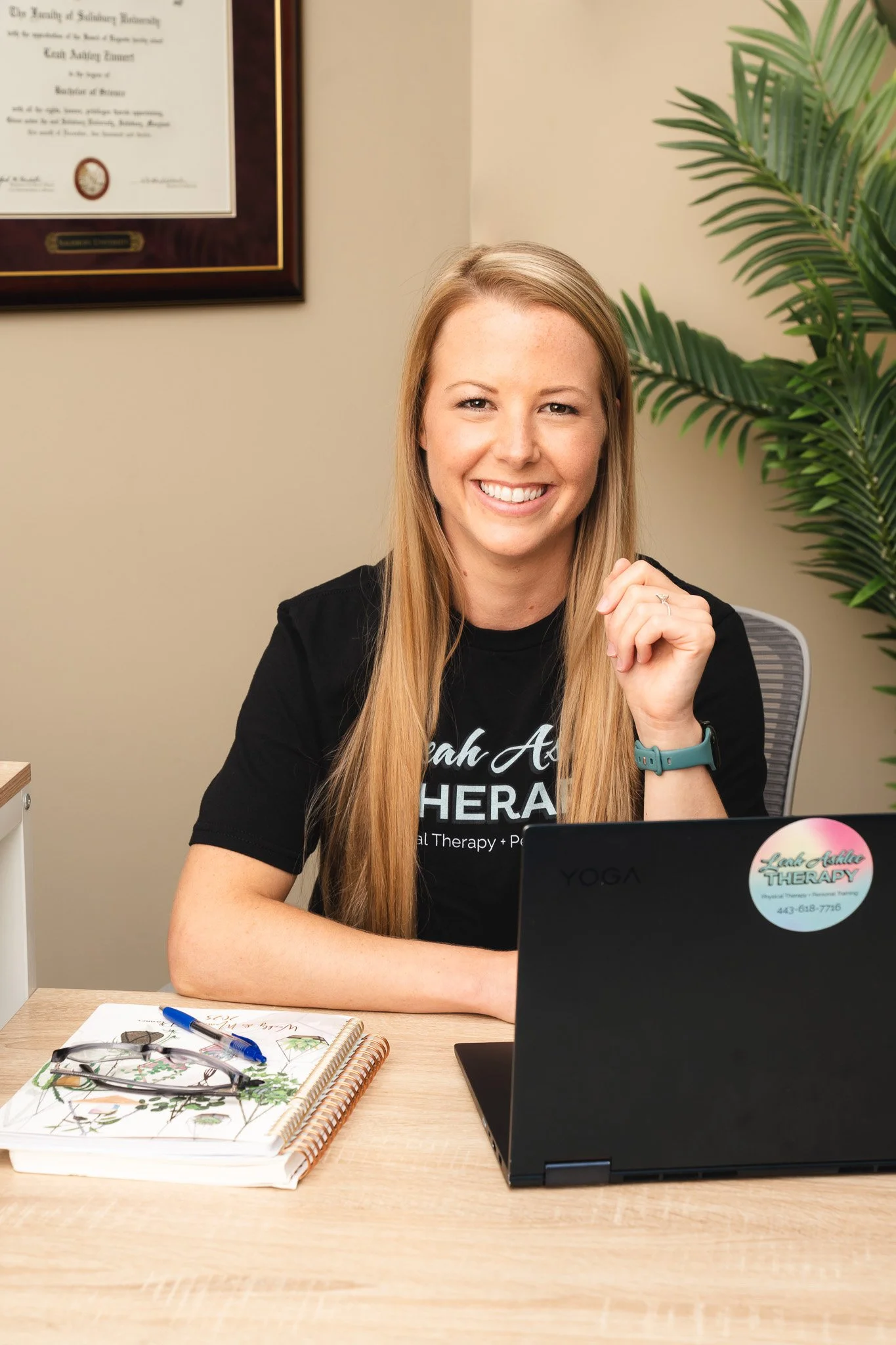 A woman smiling at a desk with a laptop, notebook, and pen, inside an office with a framed certificate on the wall and a large green plant.