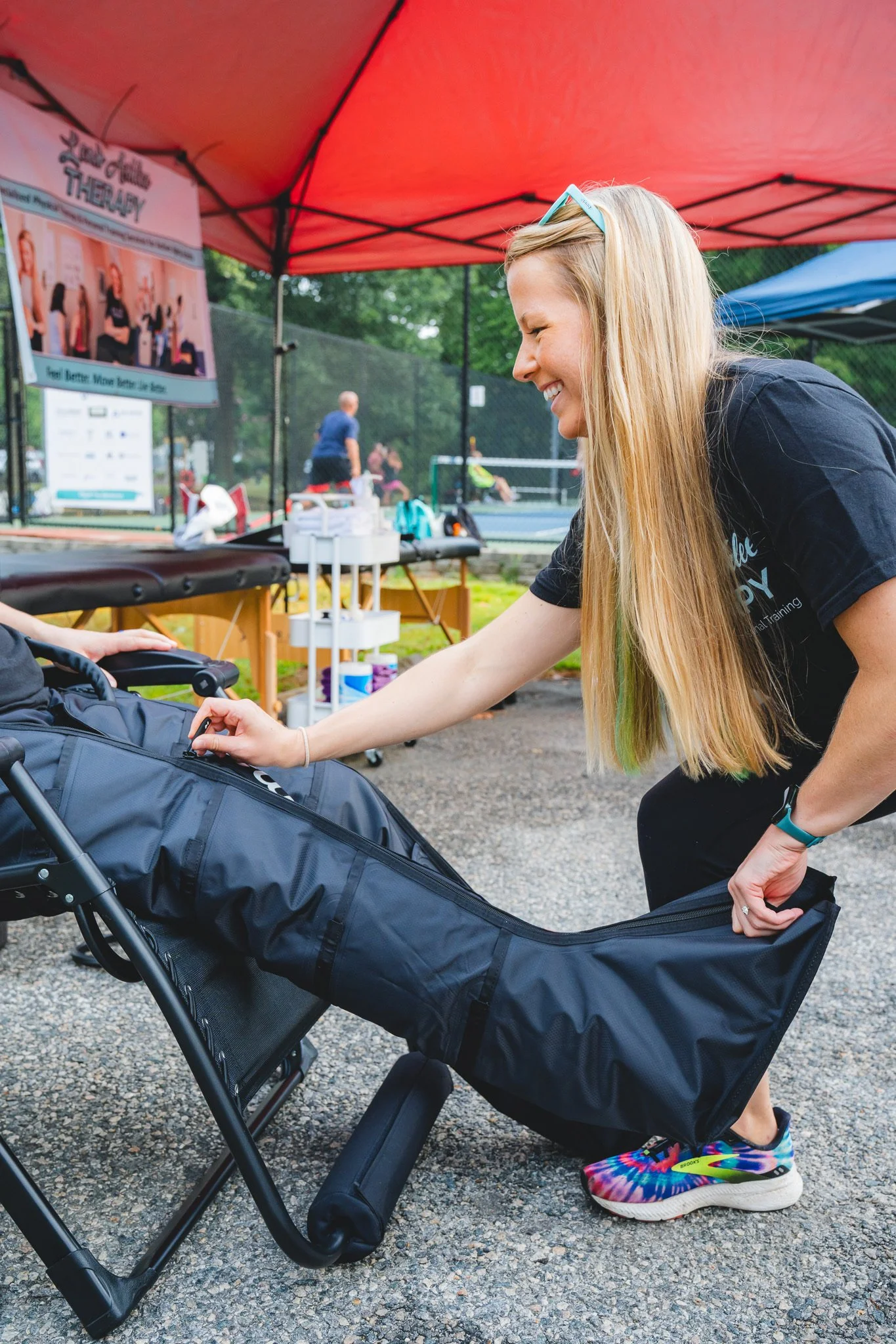 A woman with long blonde hair, wearing a black shirt and colorful sneakers, smiling and leaning over a black therapy bed outside, under a red canopy tent, with tennis courts and people playing in the background.