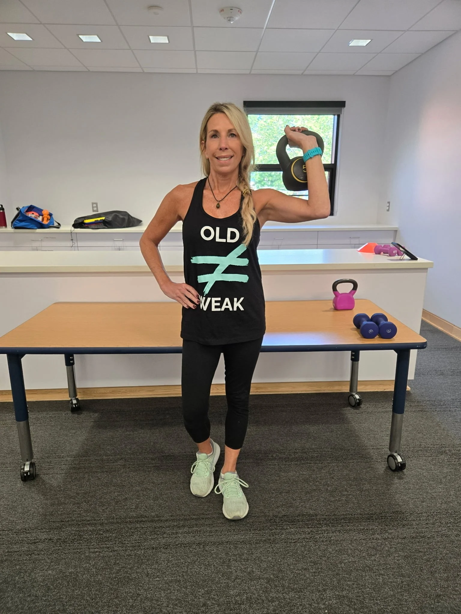 A woman working out in a fitness room with a kettlebell in one hand and a watch on her wrist. She is wearing a tank top that says 'OLD ≠ WEAK', black leggings, and sneakers.