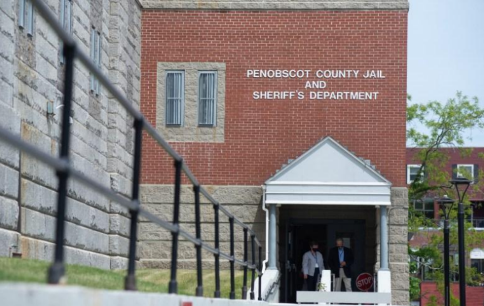 The Penobscot County Jail and Sheriff's Department building with two people standing at the entrance, a stone and brick structure, with a brick wall and a black fence in the foreground.